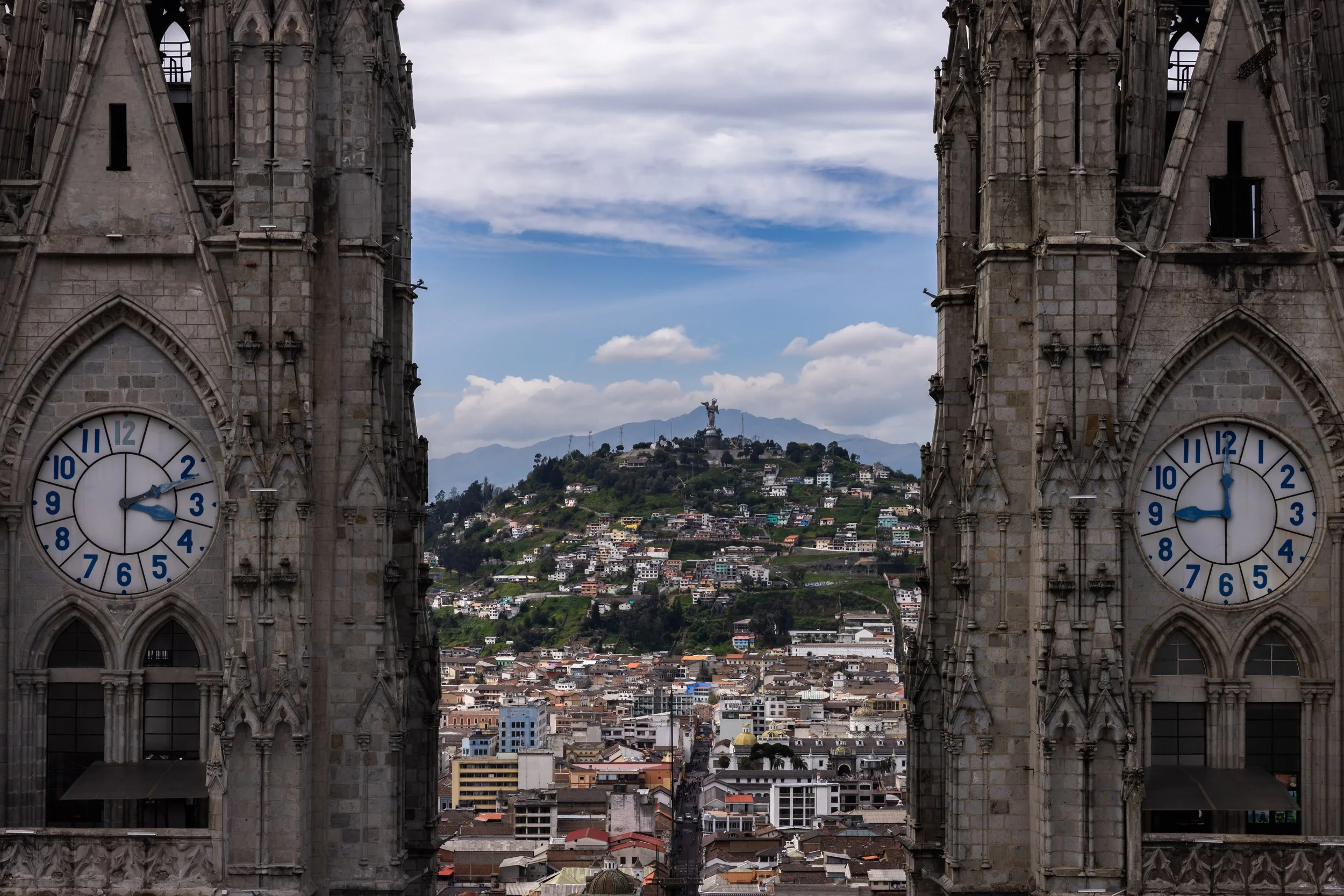 View of a cityscape through the Gothic-style windows of a cathedral, featuring two clock towers with blue clock faces and a distant hill with a statue on top.