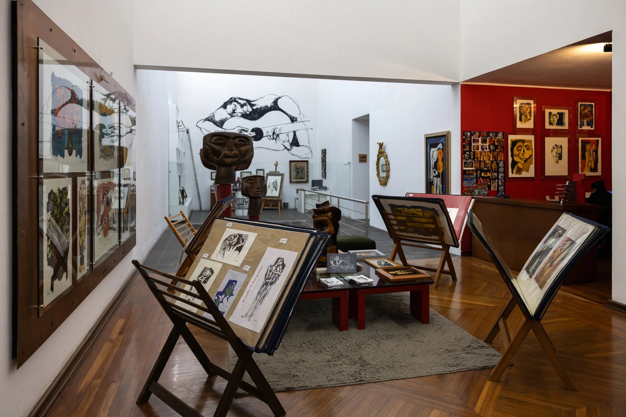 An art gallery with various artworks on display. The front features tables with drawings and sketches, including portraits and figure studies. The left wall has framed paintings and sketches, and a large black and white mural of a person playing guit