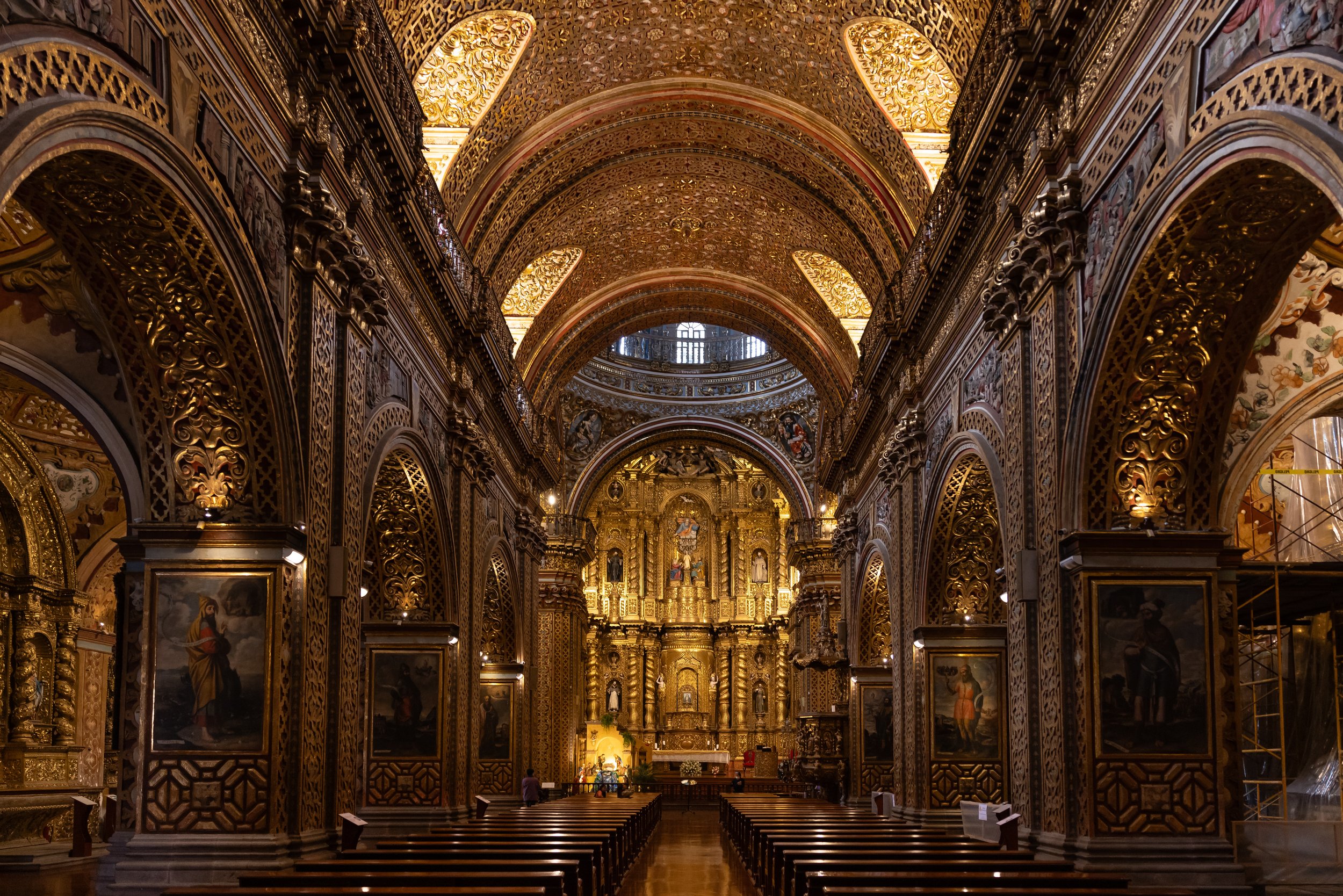 Interior of an ornate church with detailed gold carvings, arched ceilings, and religious artwork, with pews lined up in rows.
