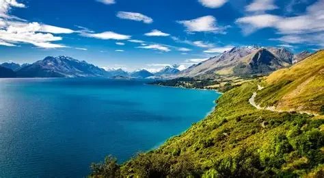 Scenic landscape with a large lake, mountains with snow-capped peaks, and a partly cloudy sky.