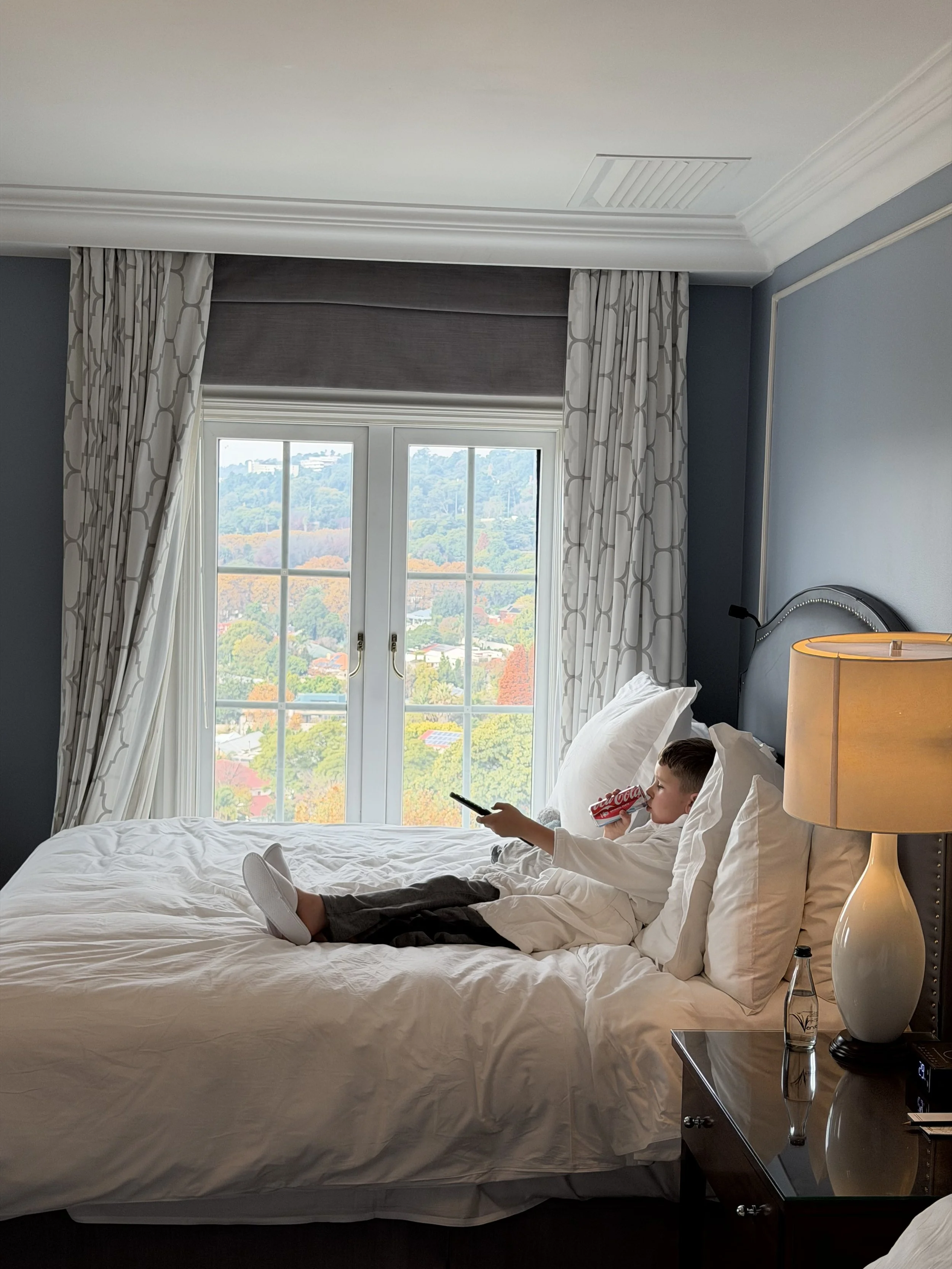 Jed lying on a bed in a hotel room, holding a smartphone in one hand and a can of Coca-Cola in the other, with a view of trees and hills outside the window.