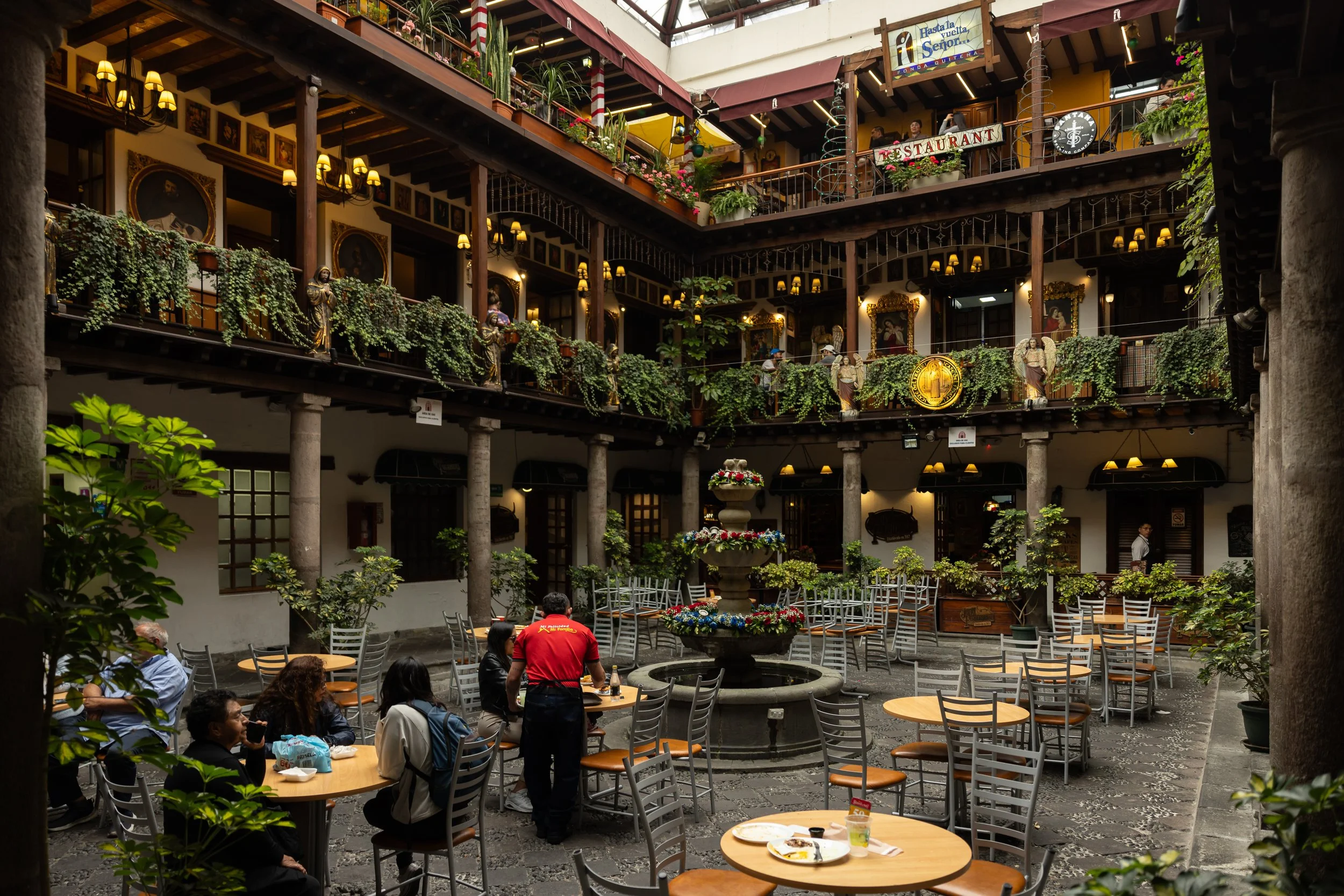 Indoor courtyard with tables and chairs, some people seated, and some standing servers serving food and drinks. The upper level has balcony railings decorated with plants and statues of saints, with various banners and signs, including a sign with th