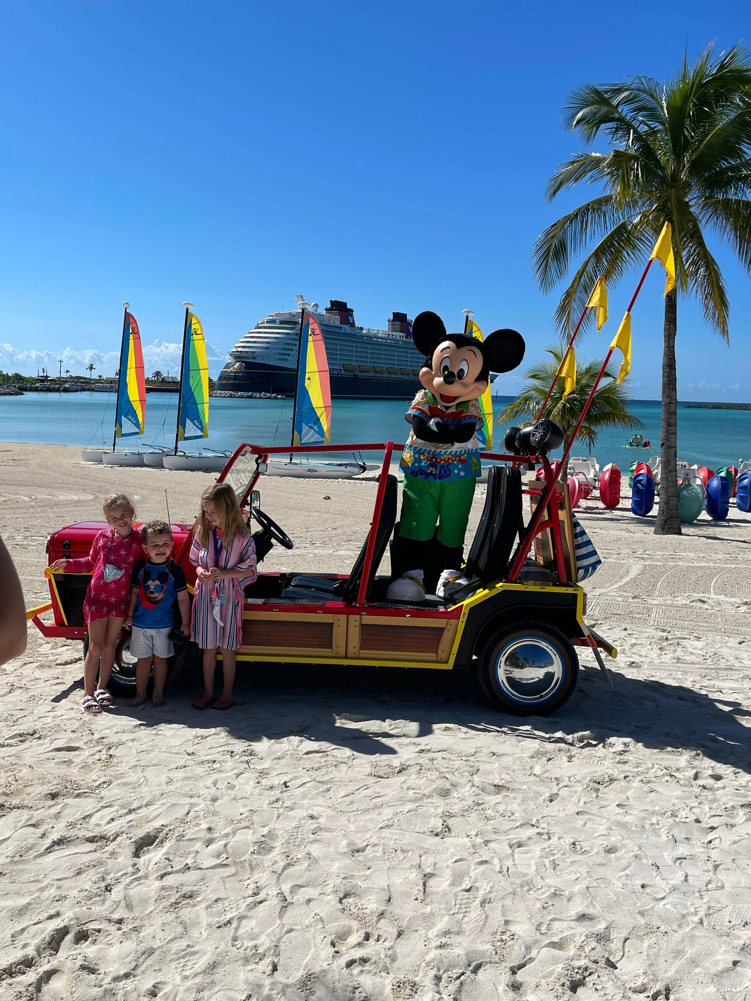 Children standing next to a vehicle with Mickey Mouse on a beach with sailboats, a large cruise ship, palm trees, and a clear blue sky in the background.