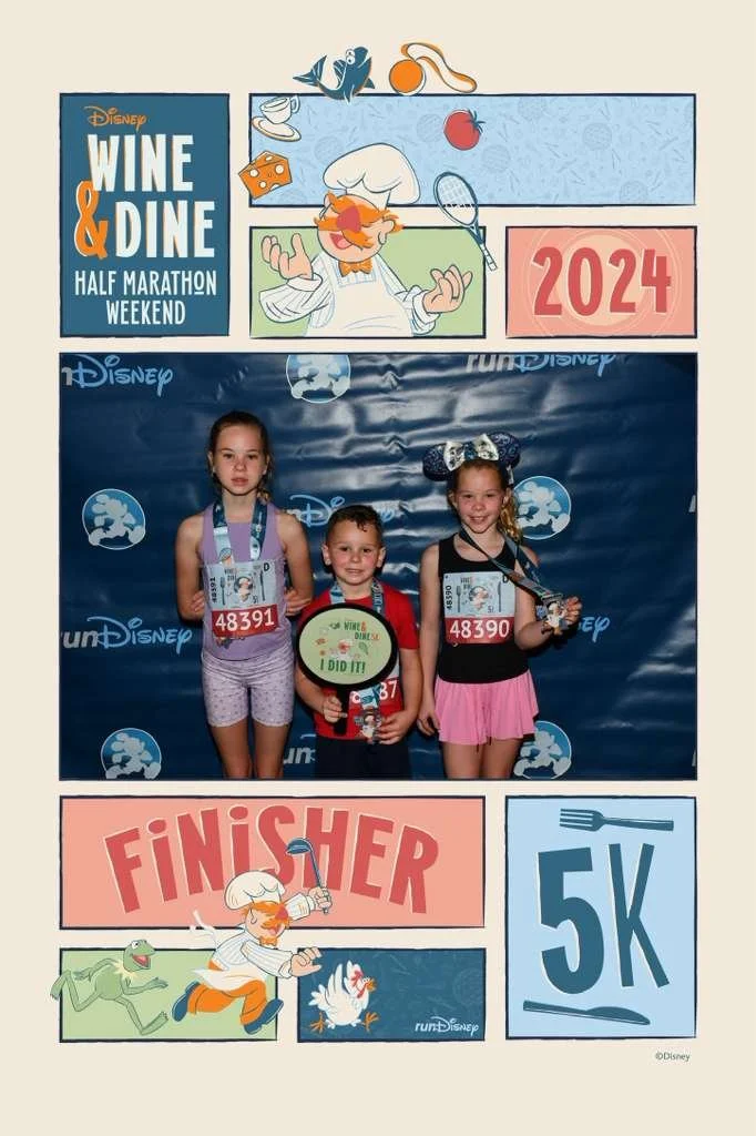 Three children at a Disney-themed 5K race wearing race bibs and medals, standing in front of a backdrop with Disney characters and race signs, celebrating their completion of the race.