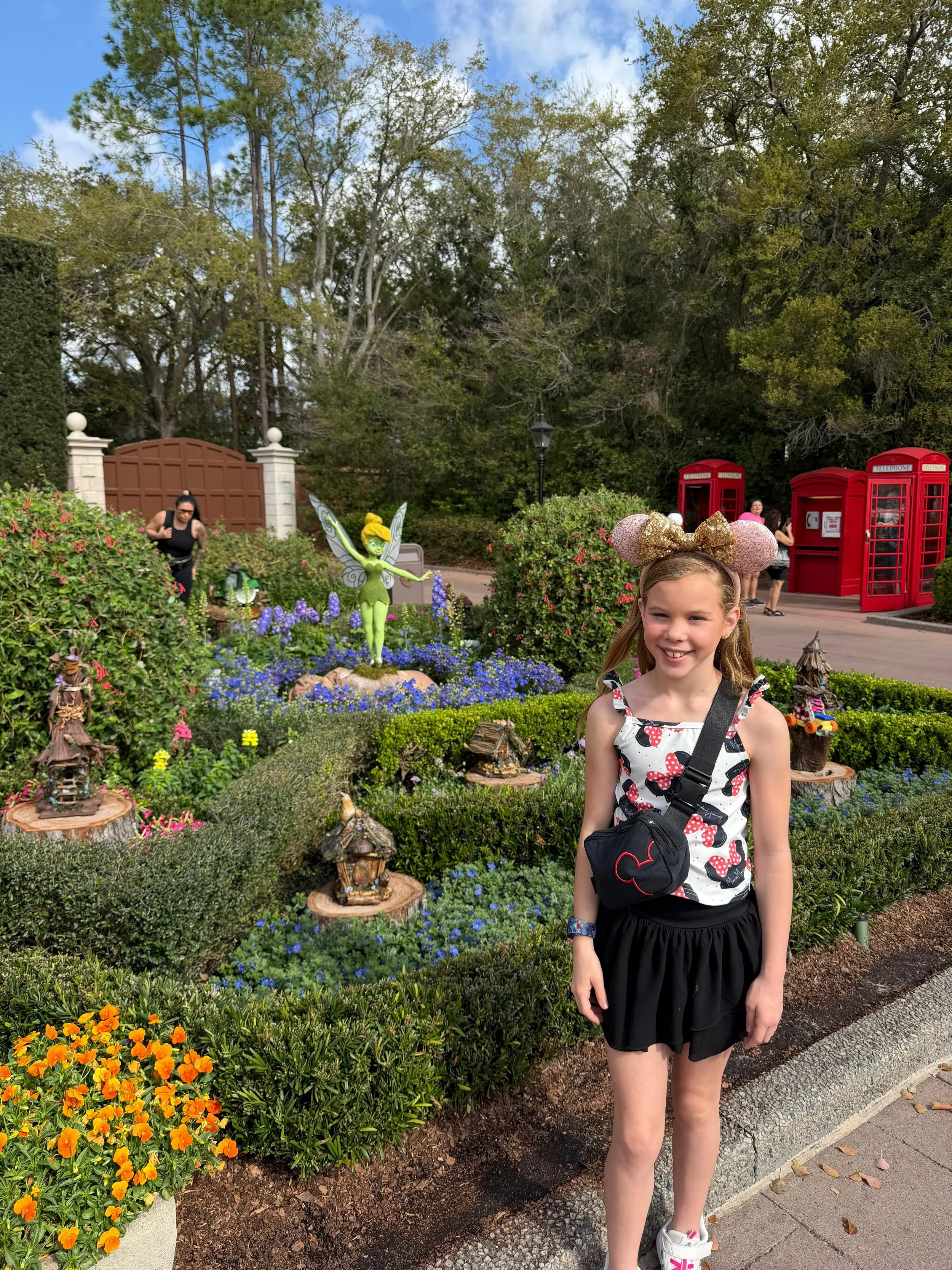 Epcot's Flower and Garden festival, posing with Tinkerbell and her houses, young girl in a Minnie Mouse tank top, Lululemon belt bag, black skort, and pink Minnie Ears