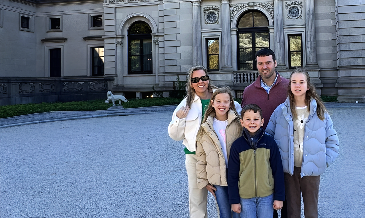 Sanders Family standing in front of a large historic building with stone architecture and arched windows. The family includes two adults and three children, all smiling and dressed in casual winter clothing.