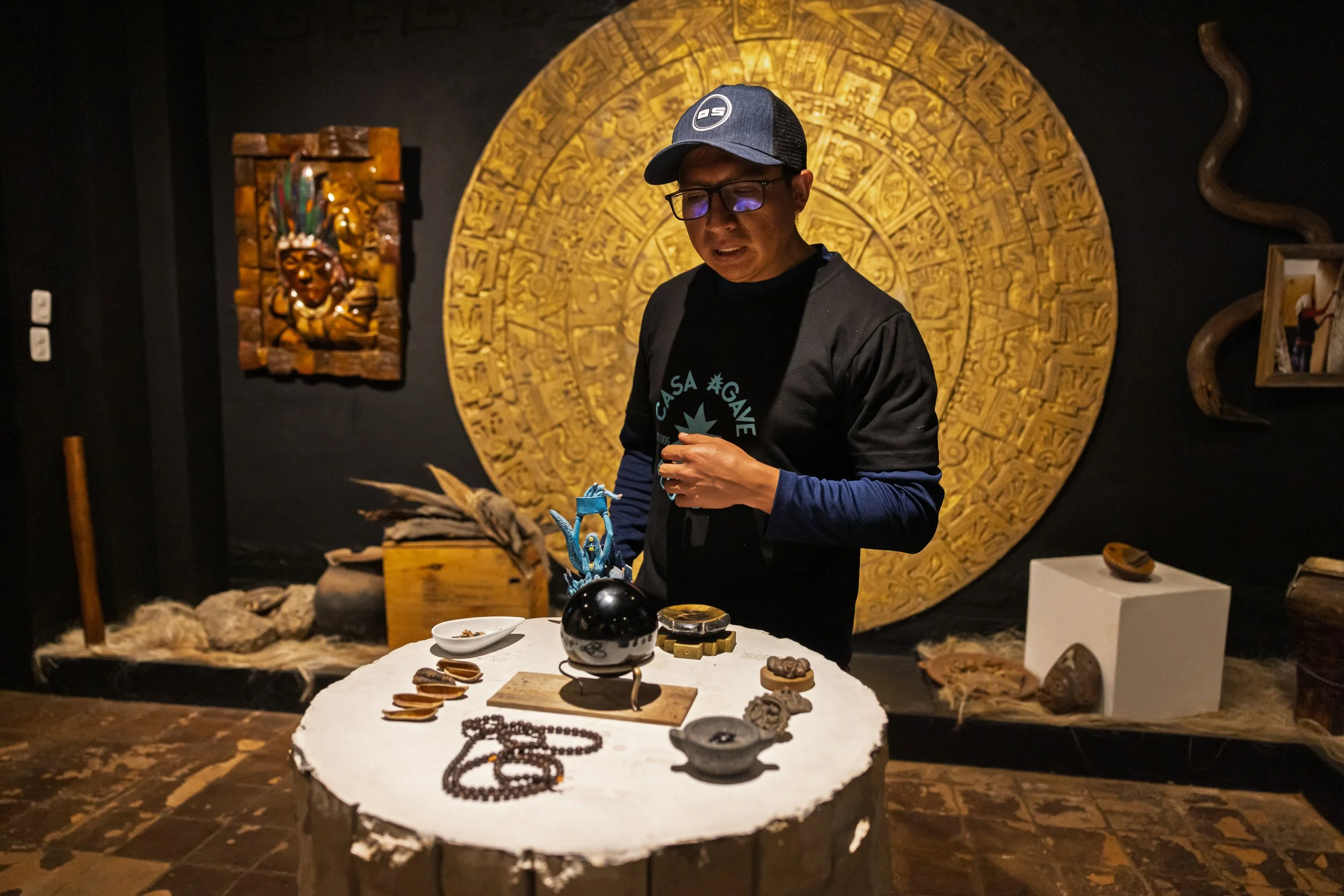 A man wearing glasses and a cap is standing behind a table with various artifacts and jewelry, in a dimly lit museum display area with indigenous art and artifacts on the wall behind him.