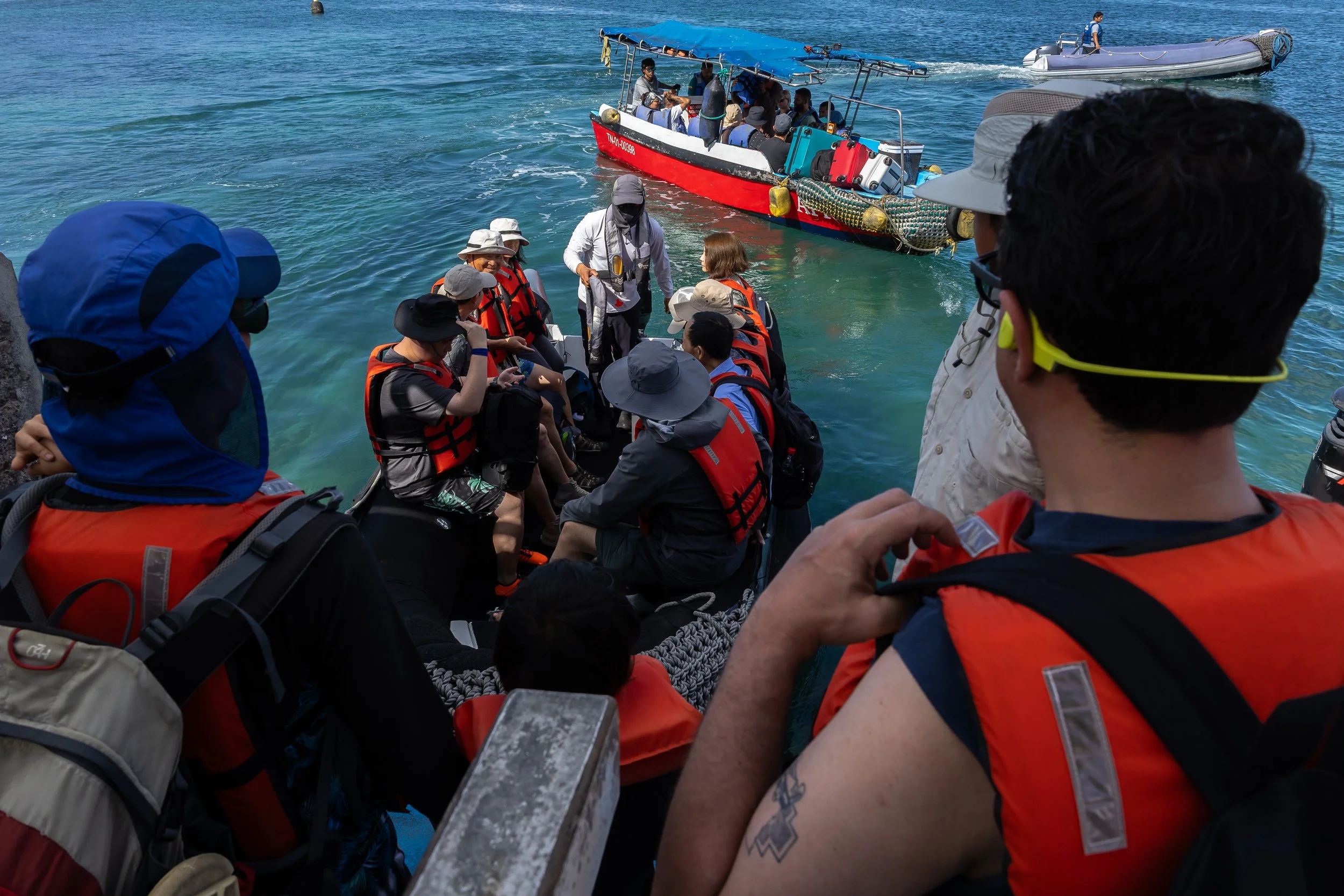 Group of people with life jackets on a boat preparing to board for a tour, with a larger boat and a smaller boat in the water.