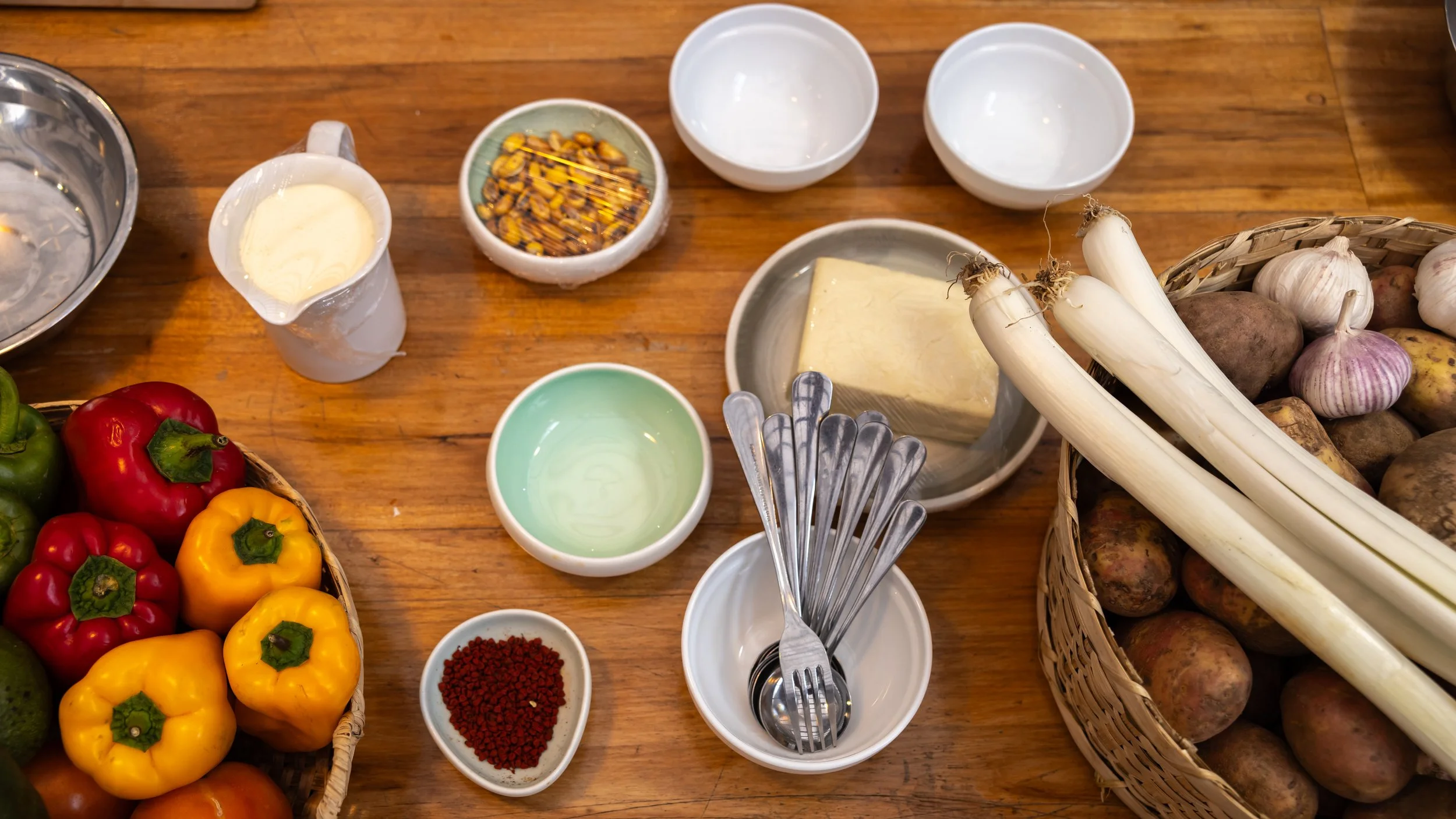 A wooden table with baskets of colorful bell peppers, potatoes, and garlic, along with bowls containing oil, herbs, and seasonings, and utensils for cooking.