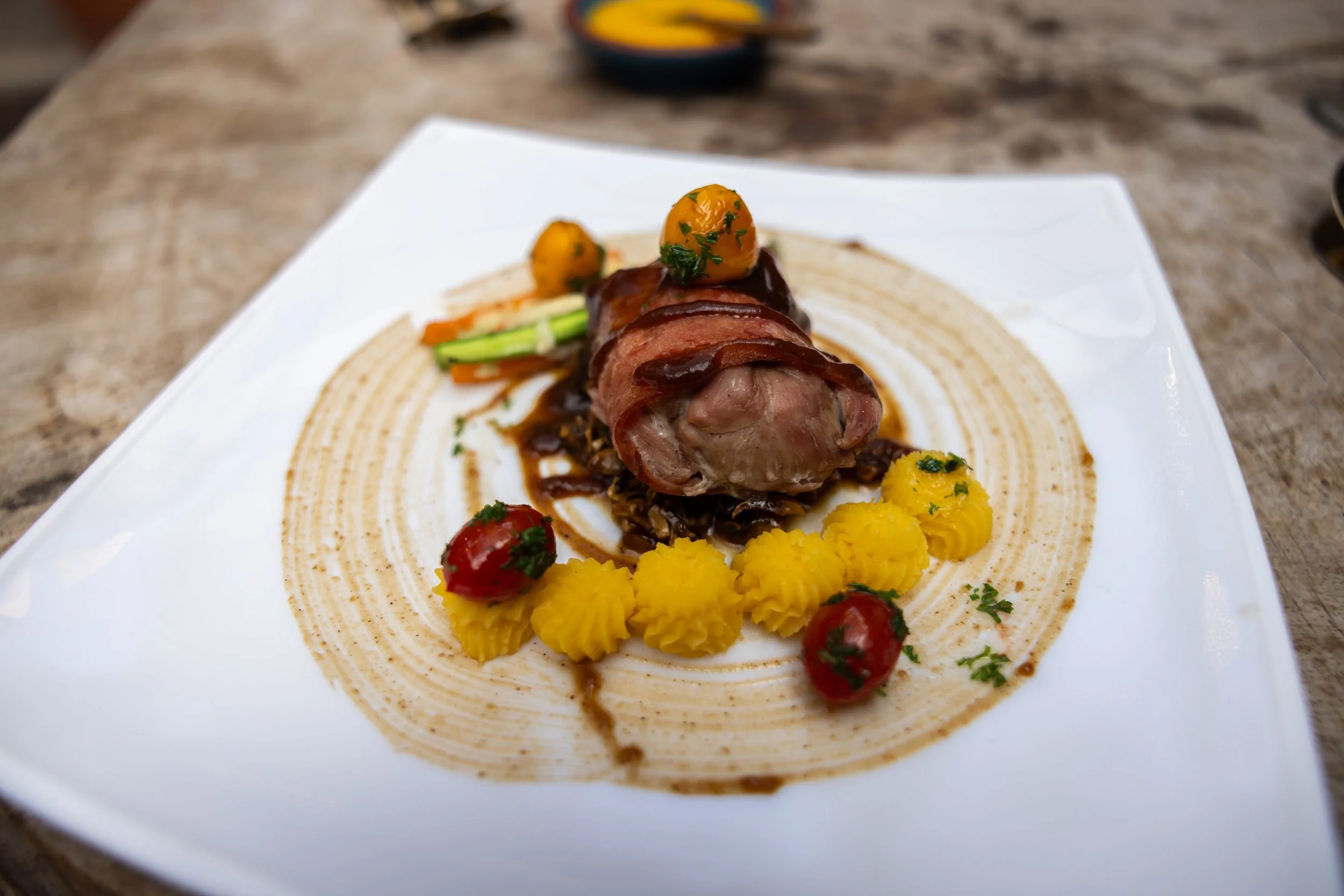 A plated gourmet dish featuring a rolled meat cut with dark sauce, garnished with small yellow potatoes, cherry tomatoes, and green vegetables, served on a white plate with a decorative pattern.