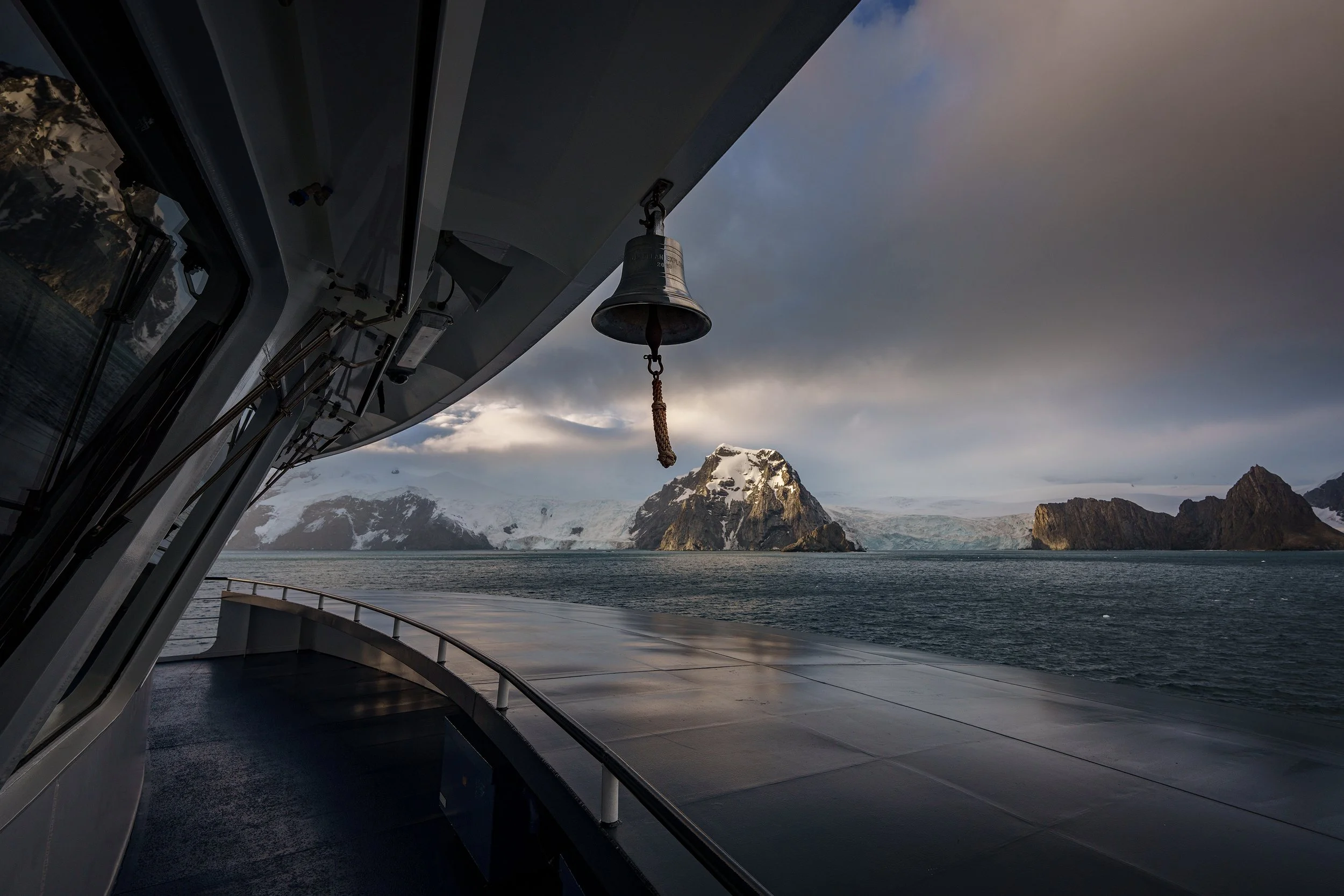 View from a boat showing a metal bell hanging from the ship's structure with a landscape of snowy mountains and rocky islands in the background under cloudy sky.