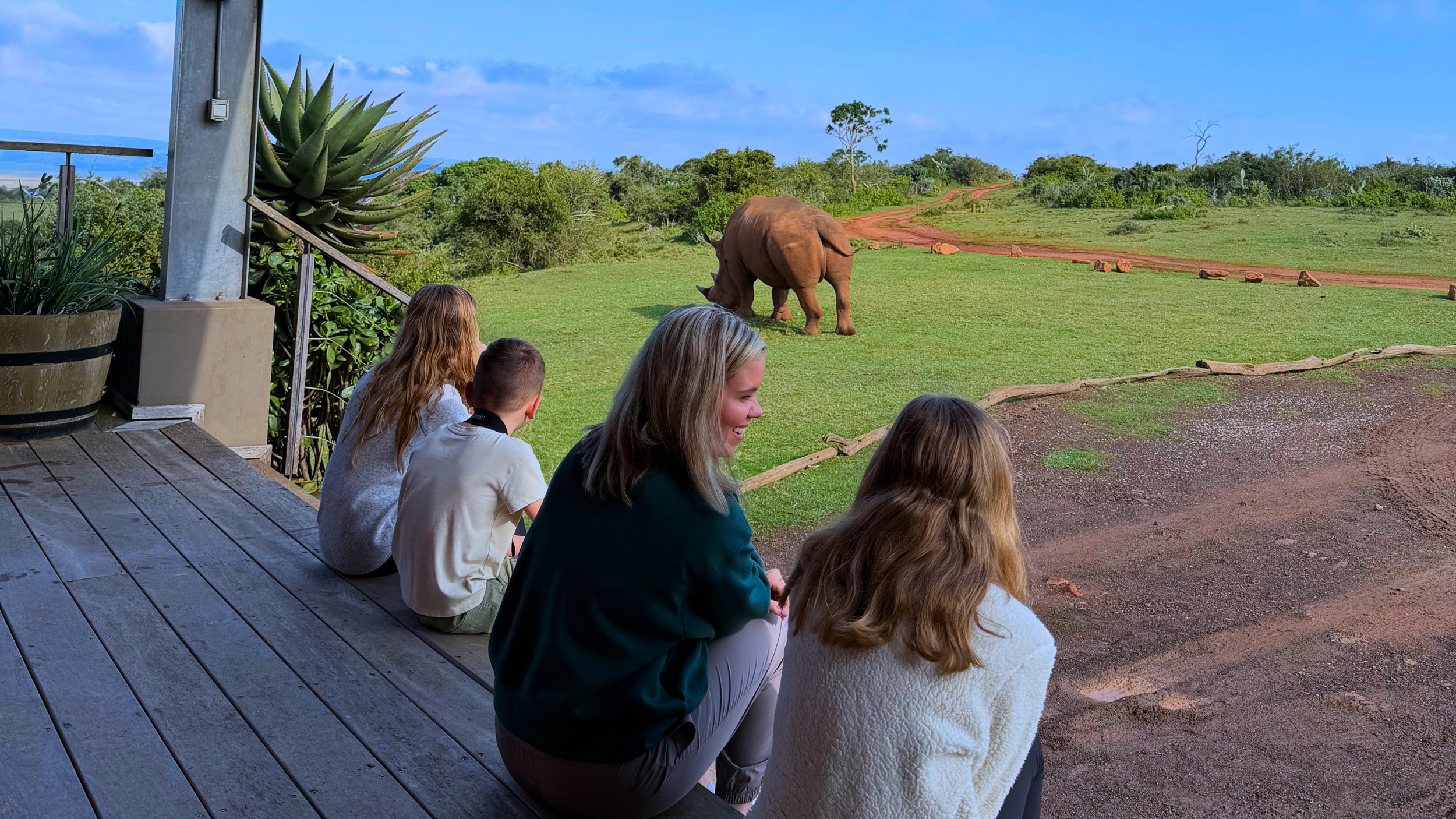 Sanders Family sitting on a wooden deck observing a rhinoceros in a green outdoor enclosure at a zoo.
