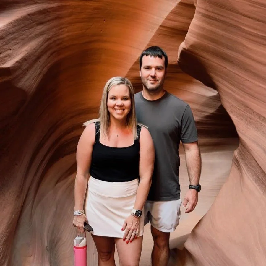 A smiling woman and man stand inside a narrow, winding canyon with smooth, reddish-brown rock walls.