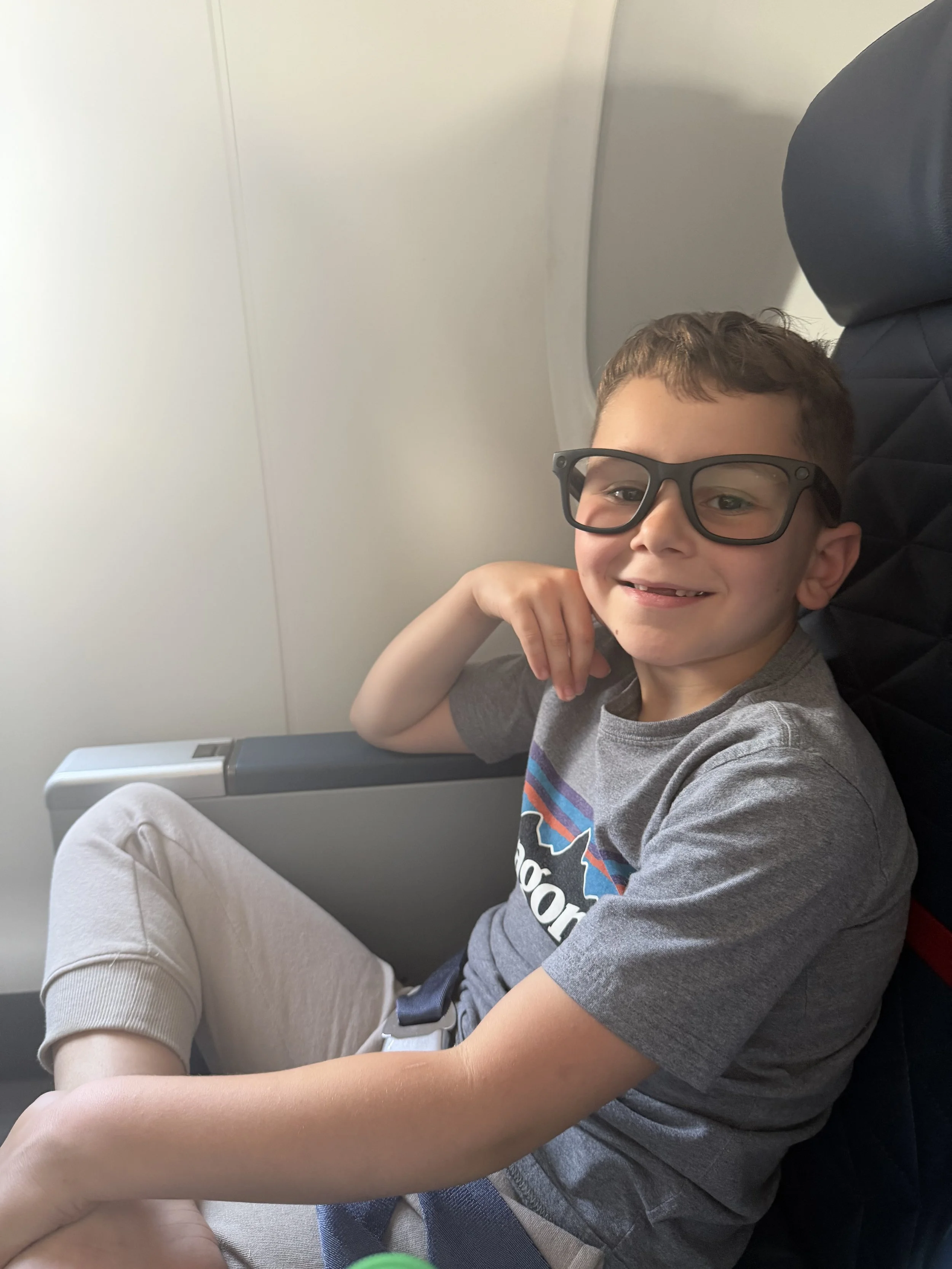 Jed with Meta glasses, smiling, sitting in an airplane seat next to the window. He is wearing a gray T-shirt and light gray pants, with his right arm resting on his lap and his left hand near his face.