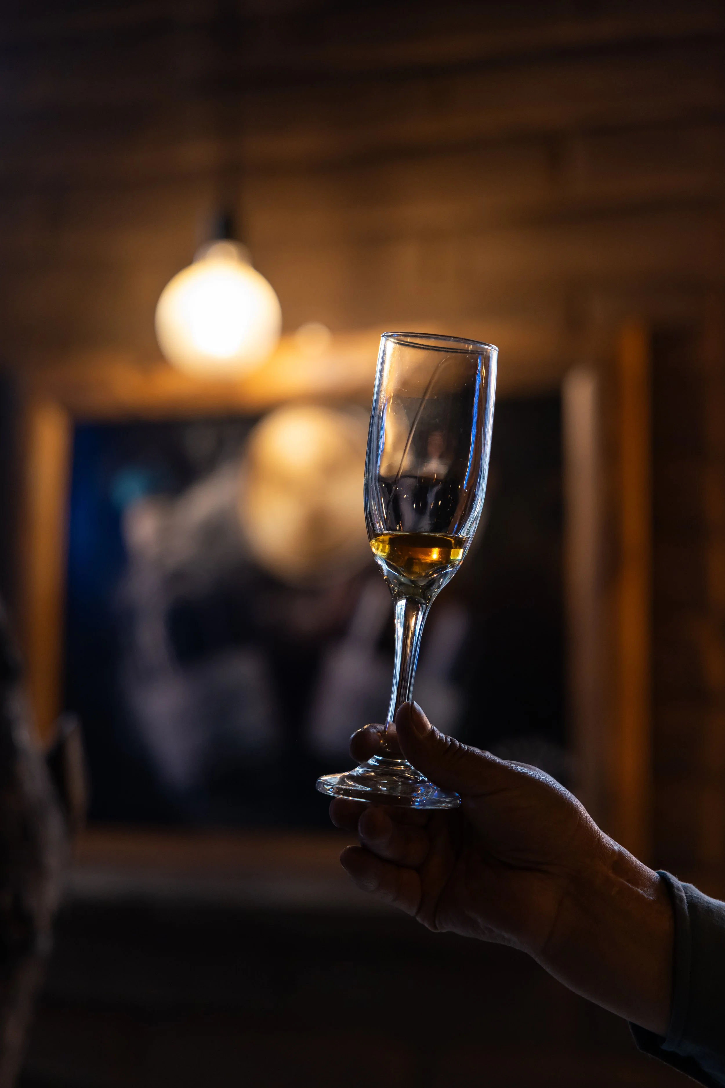 A person holding a glass of champagne with a blurred background of warm lighting and a wooden ceiling.