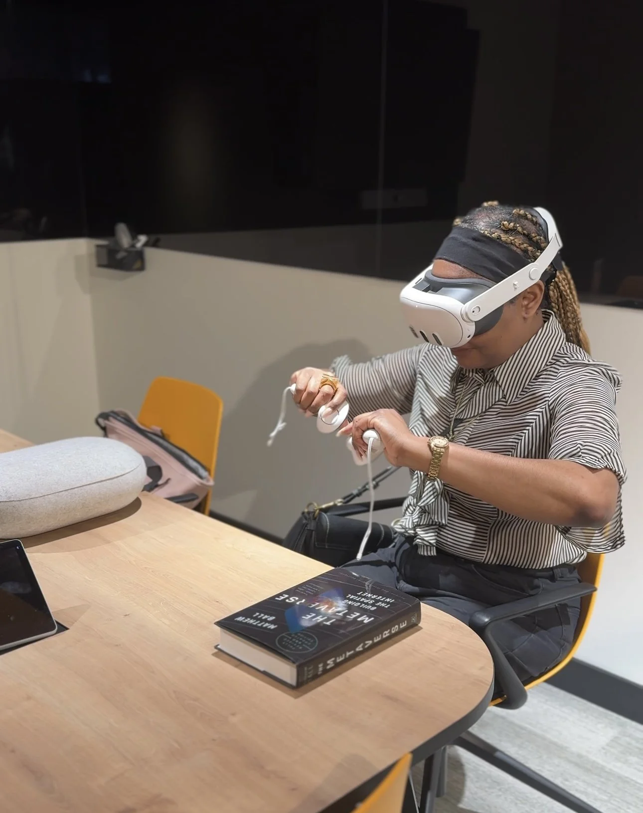 A woman wearing a virtual reality headset and headphones, sitting at a table with books, a bag, and electronic devices, in a modern office or conference room.