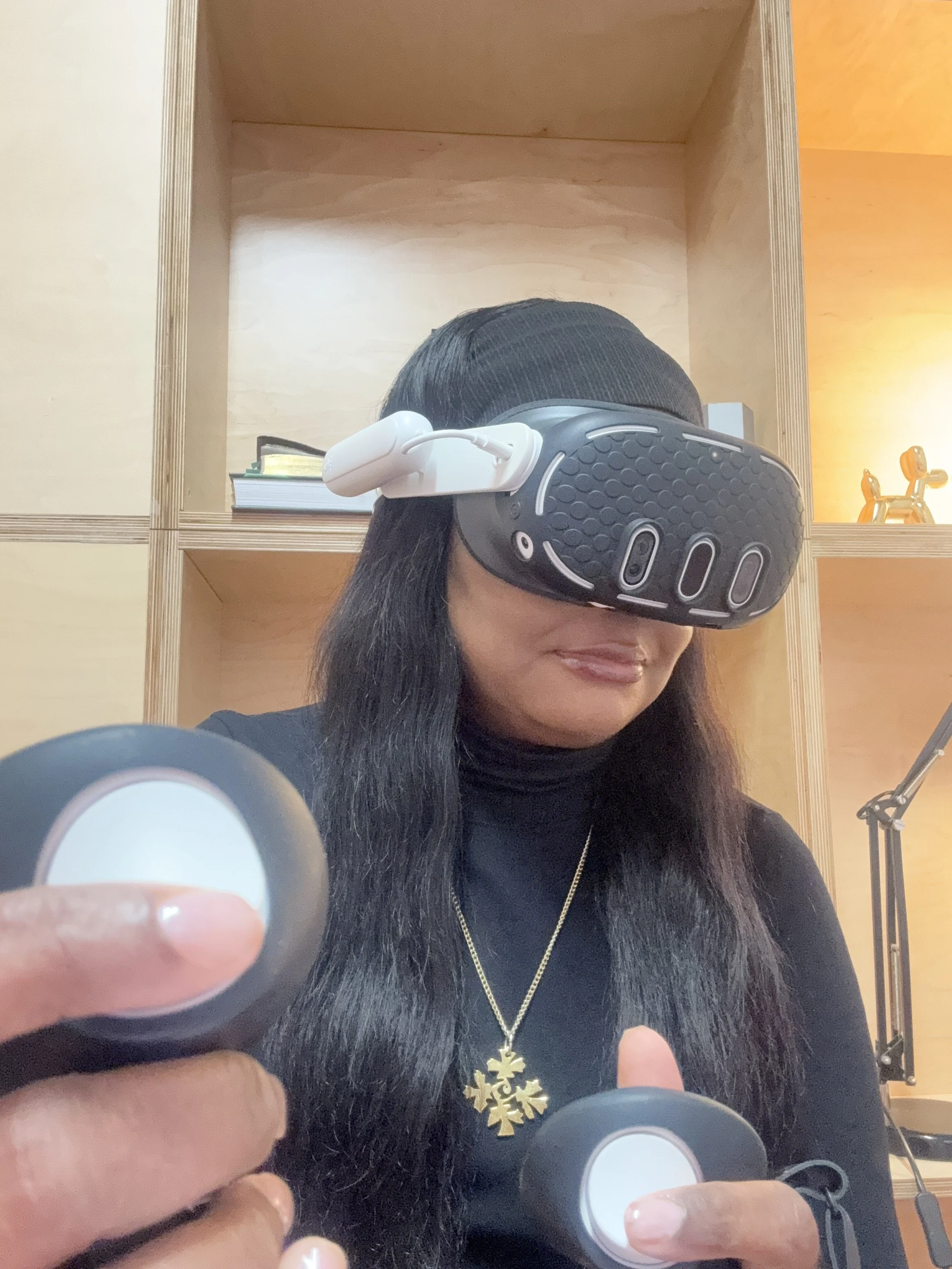 A woman with long dark hair is wearing virtual reality goggles, standing in front of a wooden shelf with books and decorative items. She is smiling and appears to be holding a controller in her hand.