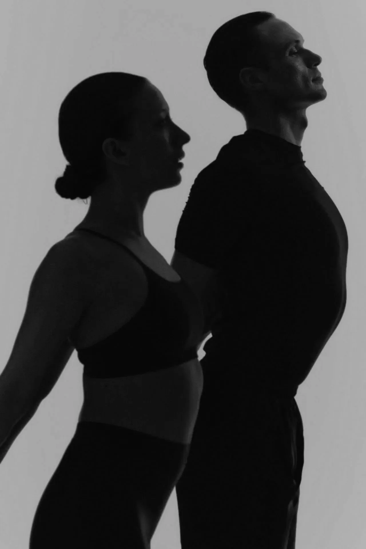 Black and white photo of a woman and a man in athletic wear, standing in profile with their backs arched. The woman on the left has her hair tied back, and the man on the right has short hair. They are positioned against a plain background.