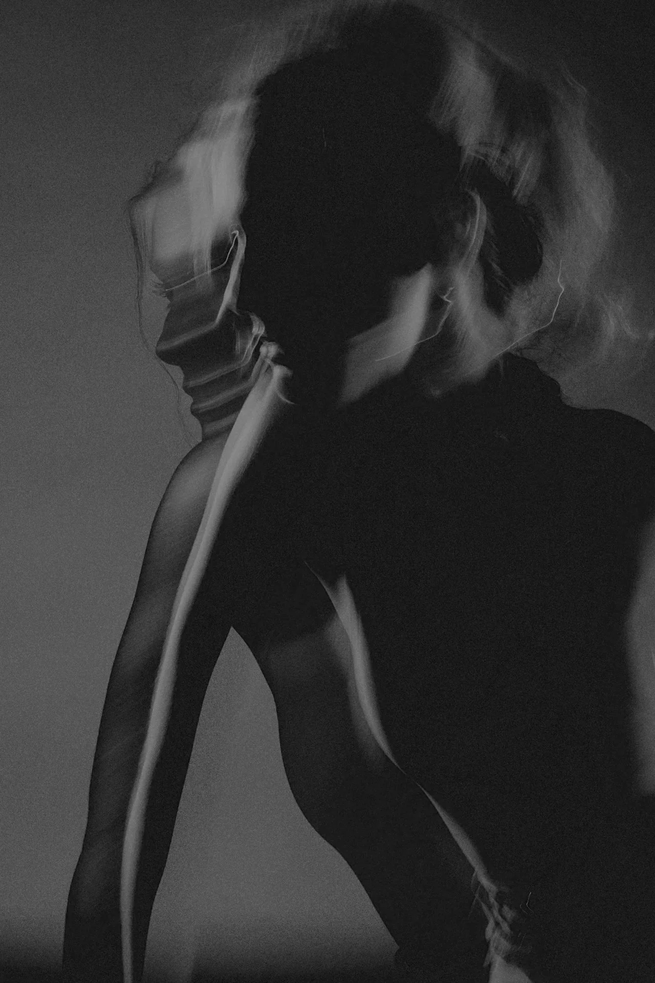 A black and white photograph of a woman with hair tied back, wearing glasses, sitting with her head resting on her hand and looking downward. The photo has a moody, artistic feel with soft lighting and shadows.