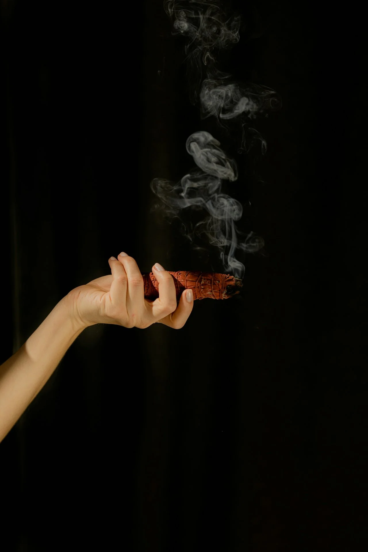 A hand holding a burning piece of wood or incense stick with smoke rising against a black background.