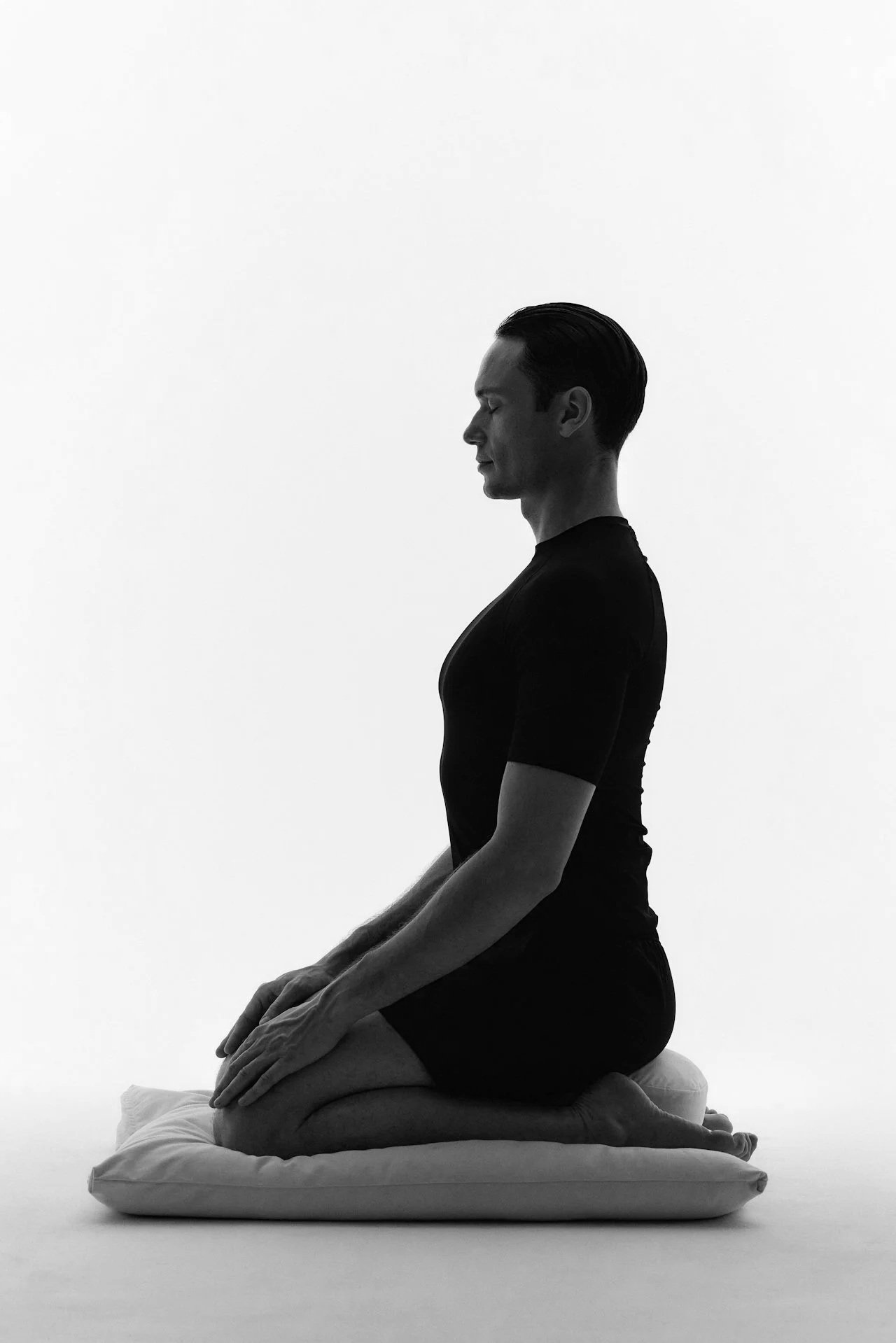 A man practicing yoga in a kneeling pose on a cushion, eyes closed, wearing a black t-shirt and shorts, against a plain white background in black and white.