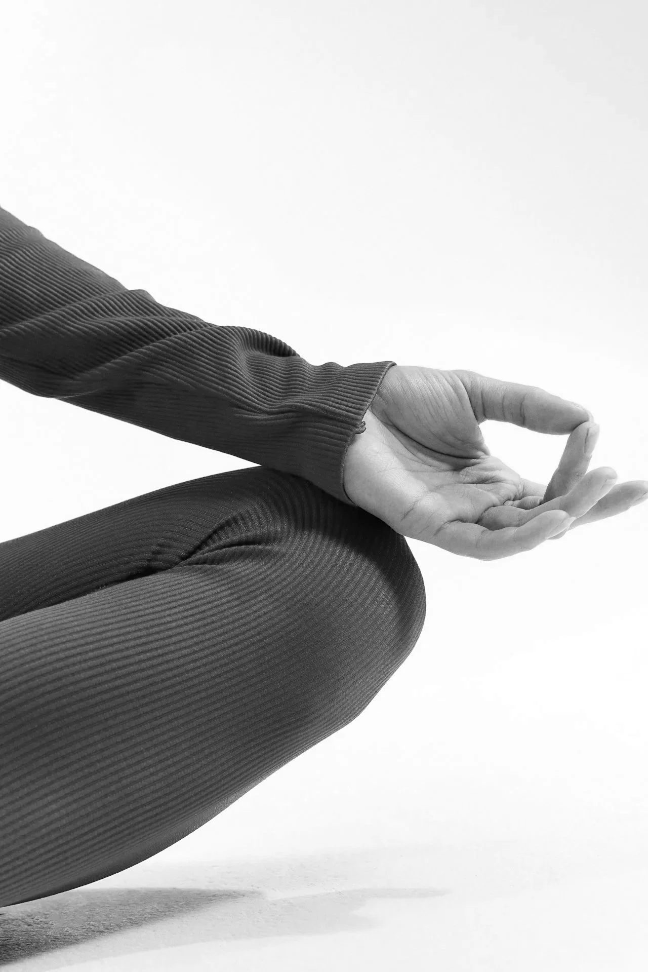 Close-up of a person performing a yoga pose, sitting cross-legged with their hand in a mudra, wearing a textured long-sleeve top and leggings, in black and white.