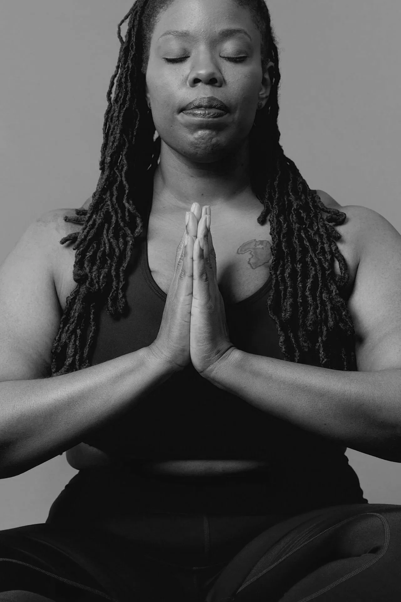 A woman with dreadlocks practicing yoga, sitting cross-legged and pressing her palms together in a prayer position with her eyes closed.