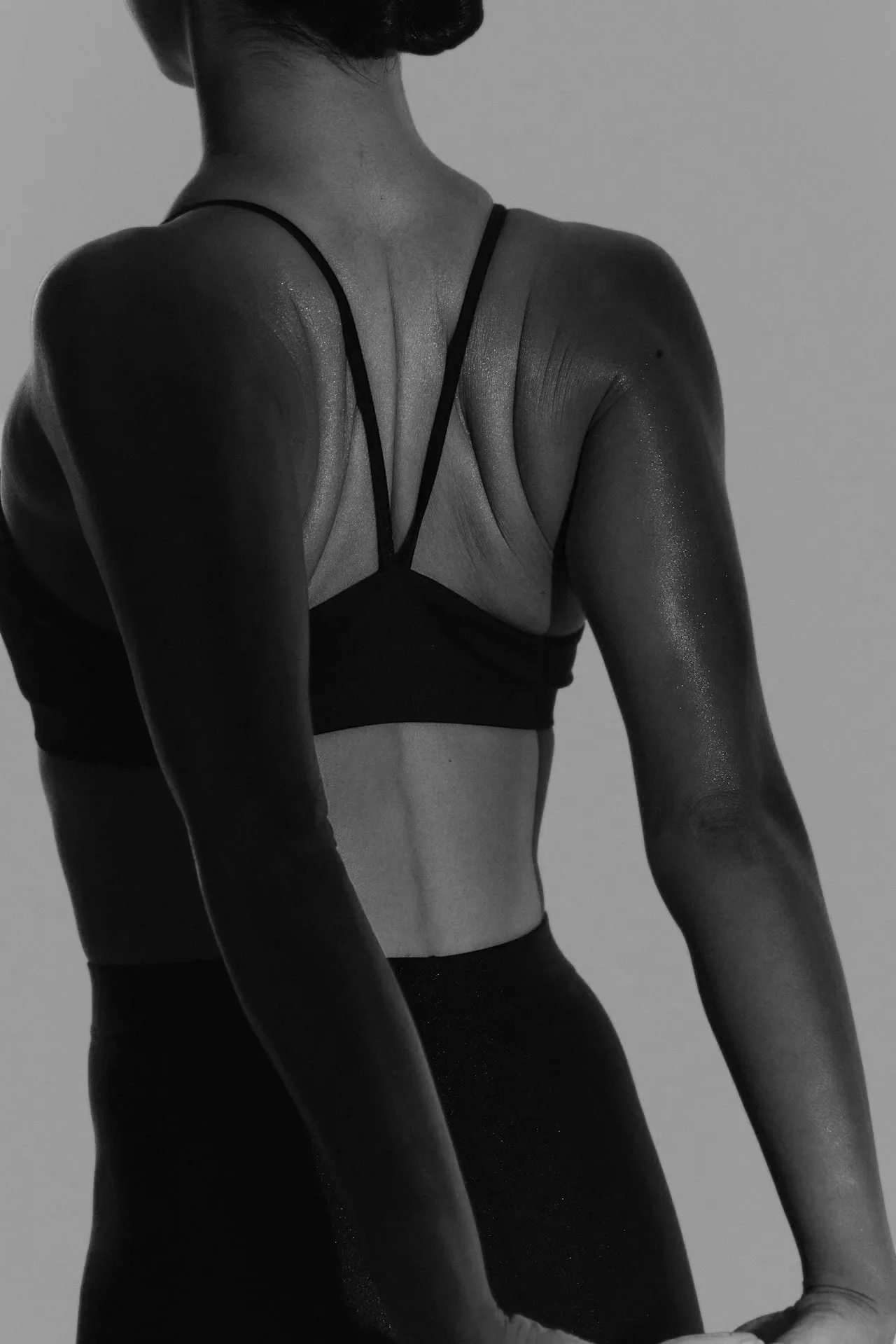 A black and white photograph of a woman from the back, wearing a sports bra and leggings, with her arms by her sides.