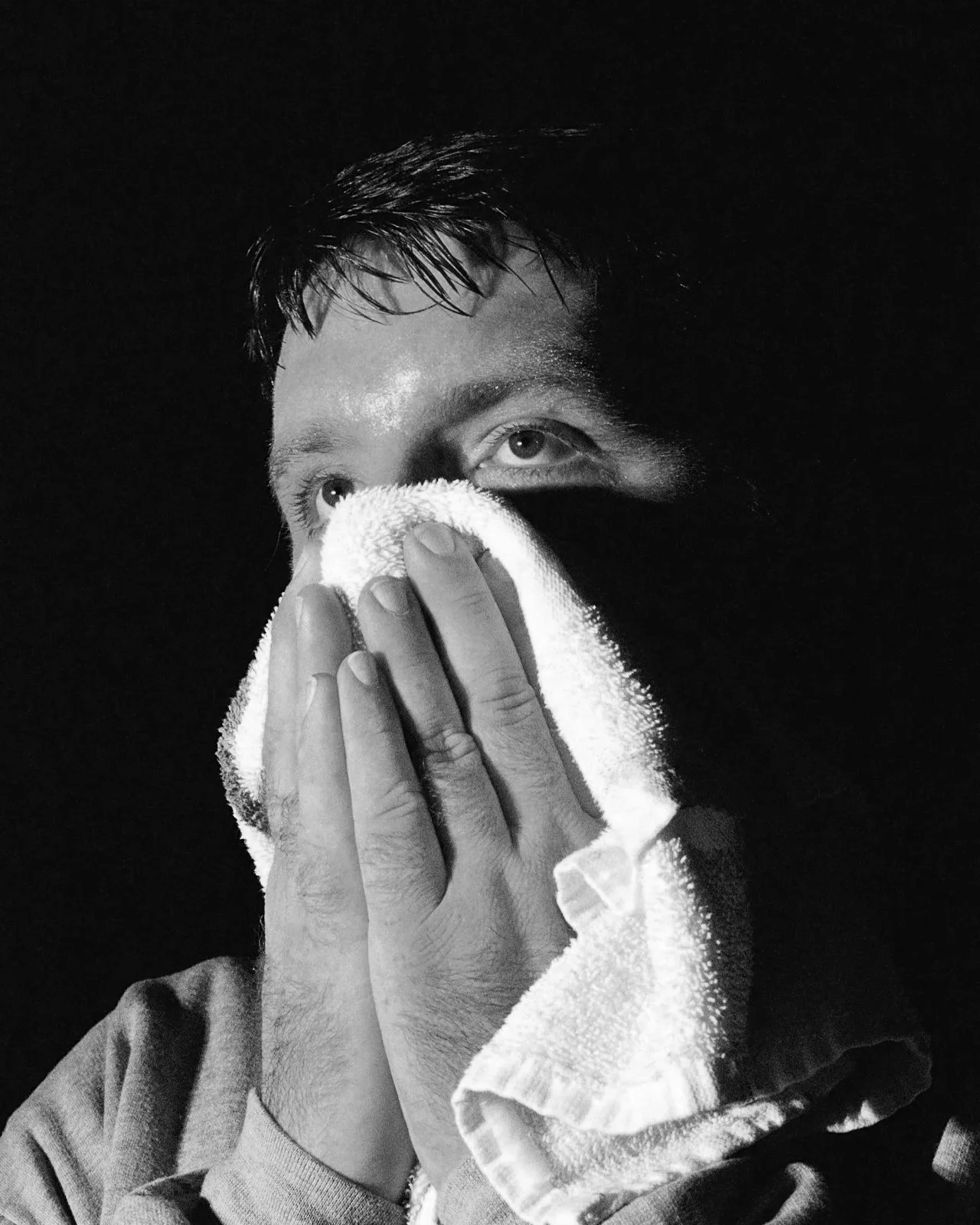 A black-and-white photo of a person with wet hair holding a towel up to their face, covering their nose and mouth, with only the eyes visible.