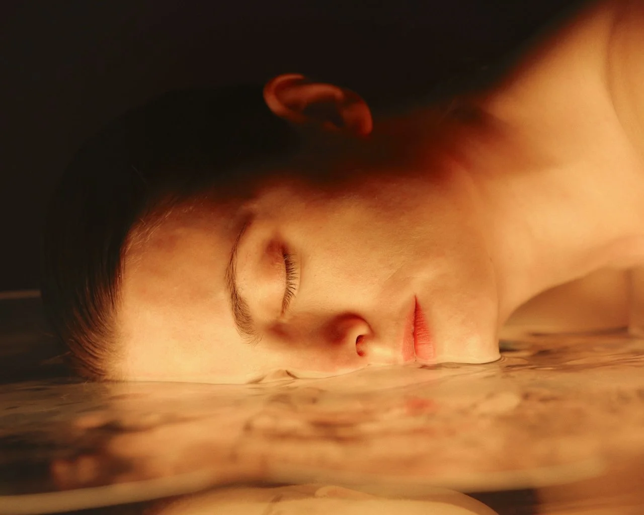A person with closed eyes submerged in water, resting their face on the water's surface.