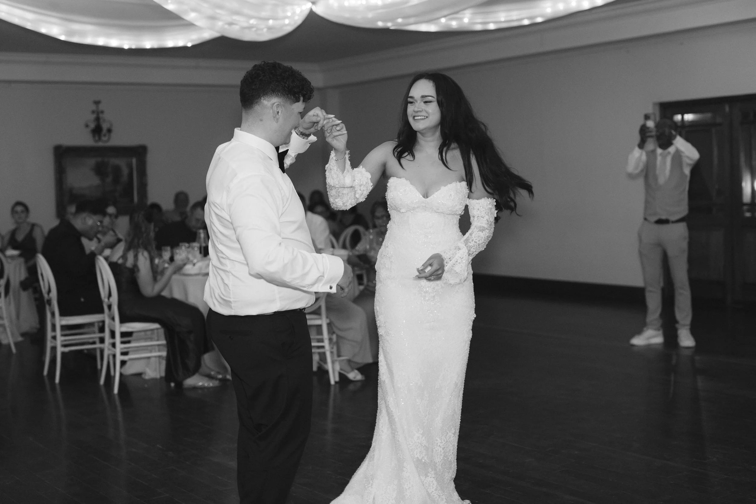 A bride and groom are dancing at their wedding reception. The bride is wearing a lace wedding gown, and the groom is dressed in a white shirt with a bow tie. Guests are sitting at tables in the background, and a photographer is taking pictures.