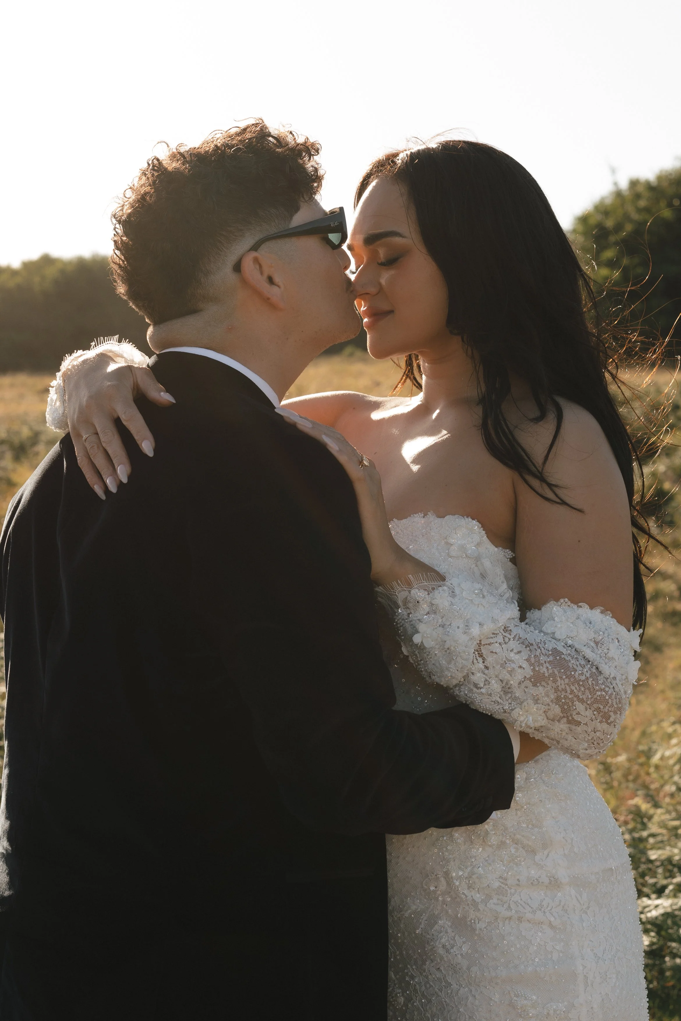 A newlywed couple stands close together outdoors at sunset, about to kiss. The groom wears a black tuxedo and glasses, and the bride wears a white lace wedding dress with off-shoulder sleeves. The background shows greenery and a clear sky. Gqeberha