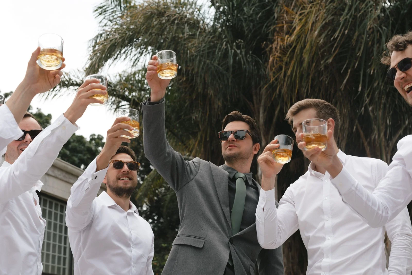 Group of men at an outdoor celebration, wearing formal and casual attire, holding glasses of whiskey and raising them in a toast, with trees and a building in the background.