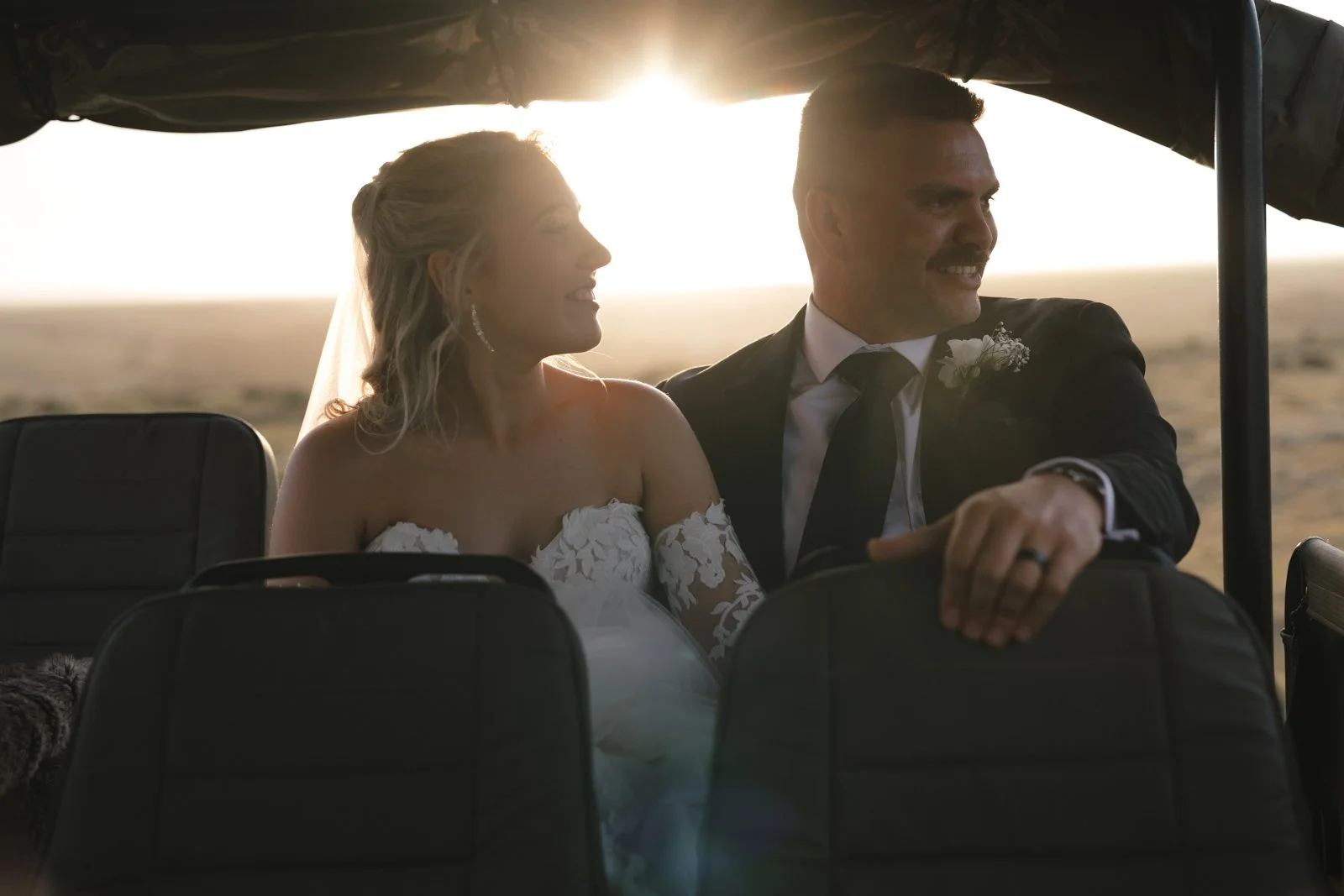 A bride and groom in wedding attire sitting inside a safari vehicle, smiling and looking at each other, with an african landscape and sunset in the background. Gqeberha wedding photographer at Lombardini Wedding Venue.