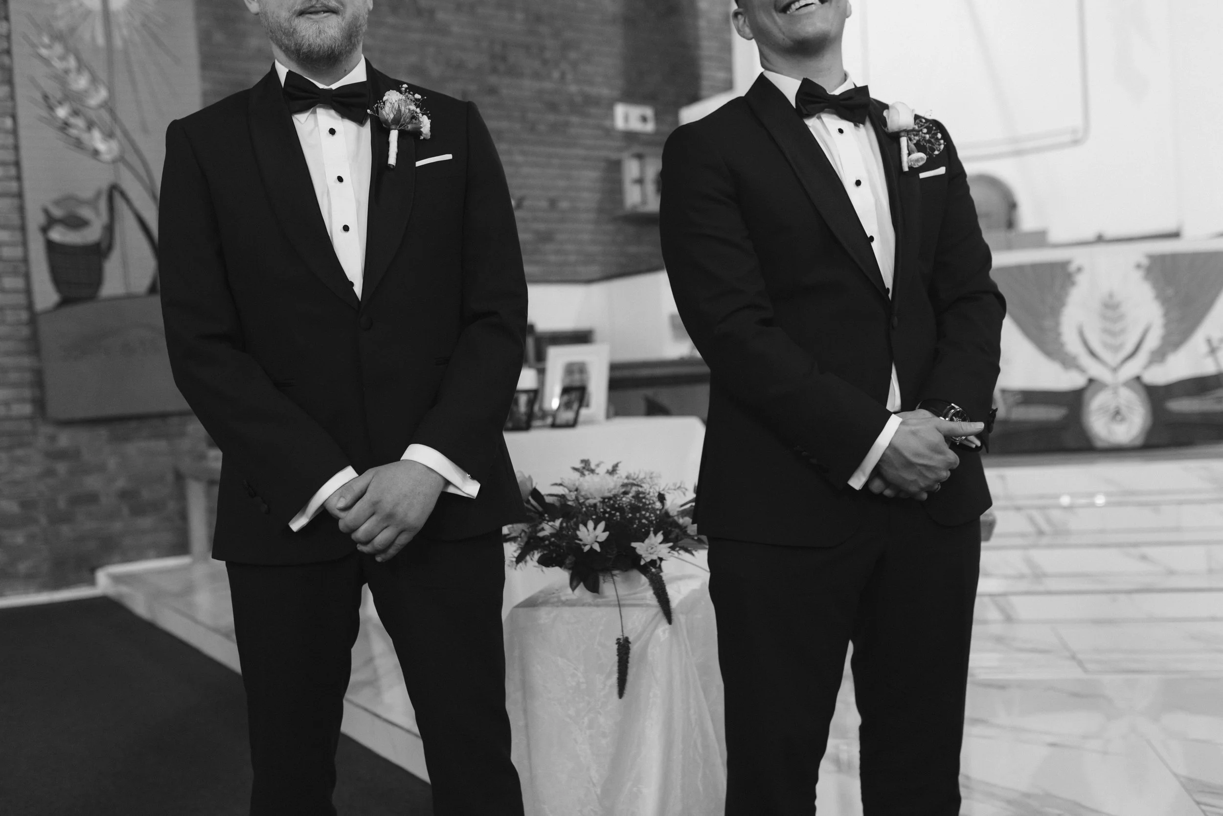 Two men in black tuxedos with bow ties standing indoors, one with a boutonnière and the other with a flower pinned to his lapel, possibly at a formal event or wedding, with a floral arrangement and a flag with an emblem in the background. Gqeberha 