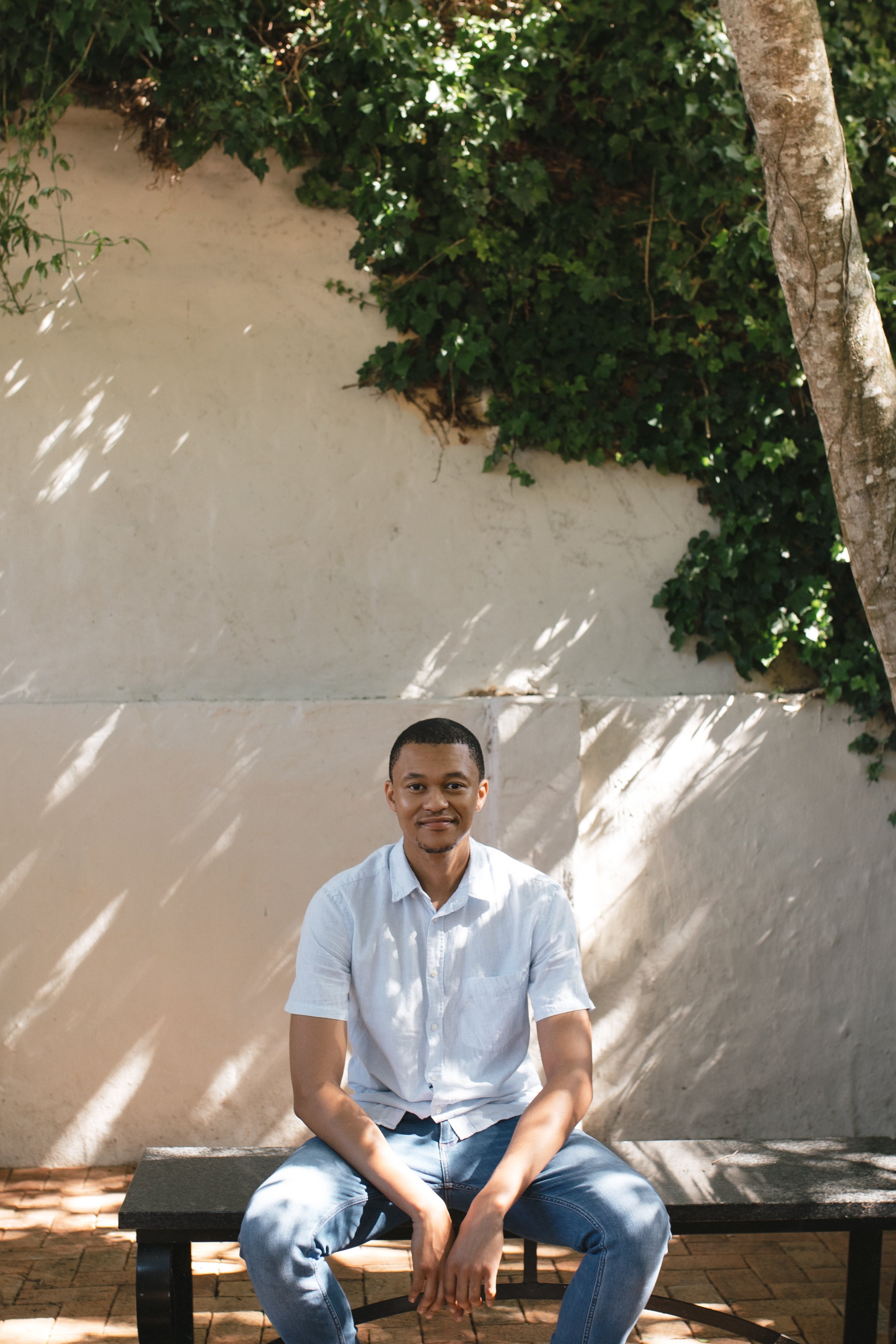 A young man sitting on a black bench outdoors, with a white wall, green leafy plants, and a tree in the background, during daylight. Gqeberha wedding photographer and videographer.