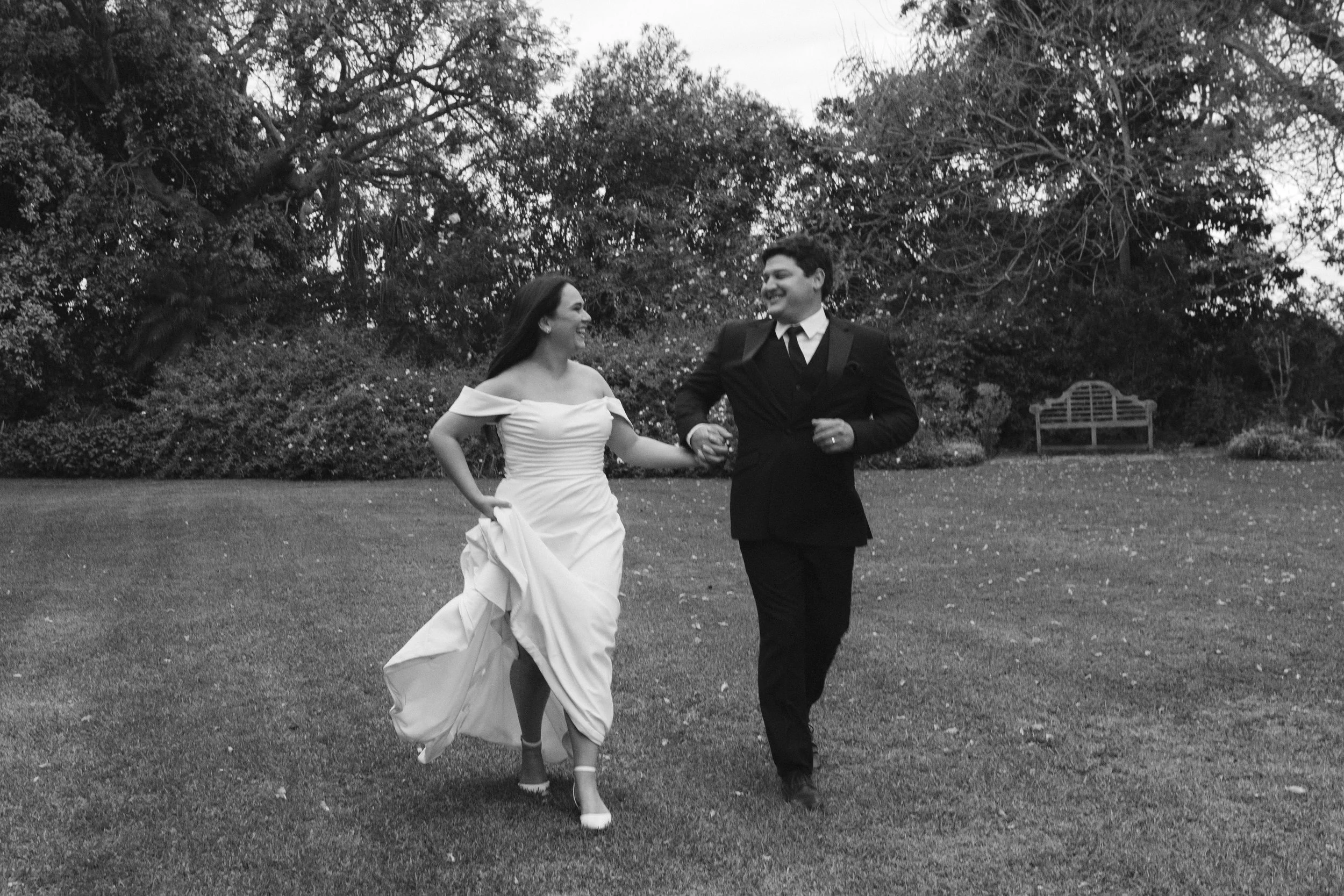 A black and white photo of a bride and groom holding hands and running outdoors on a grassy area with trees and a bench in the background. Gqeberha wedding photographer at Annerkant Wedding Venue in Kirkwood.
