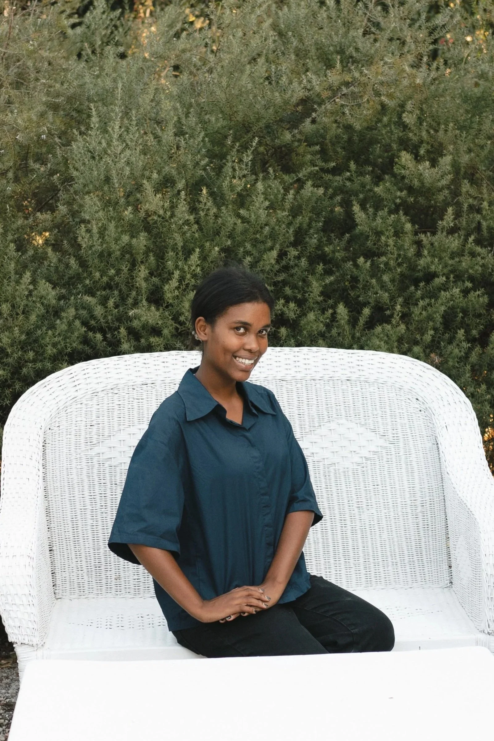 A woman sitting on a white wicker outdoor bench in front of green shrubbery, smiling at the camera. Gqeberha wedding photographer.