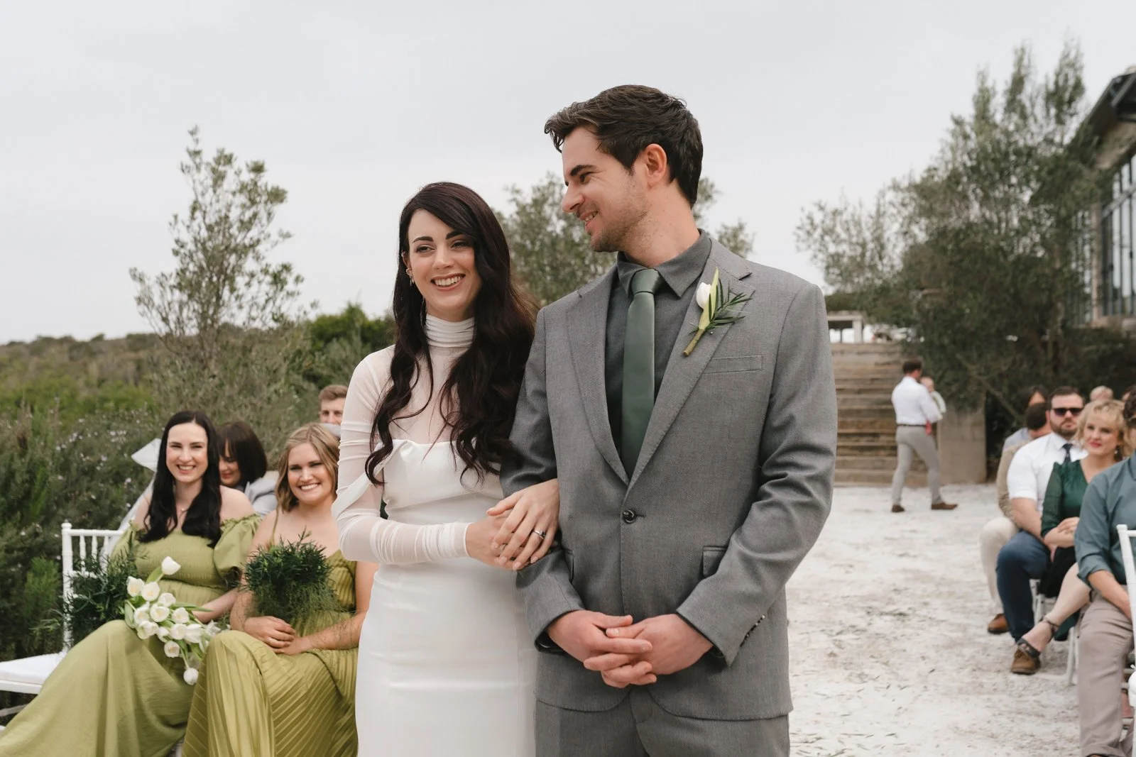 A smiling bride and groom holding hands during their wedding ceremony outdoors, with bridesmaids and guests seated in the background. Gqeberha wedding photography at The Rose Barn, Jefferys Bay, dress by Werner Dey