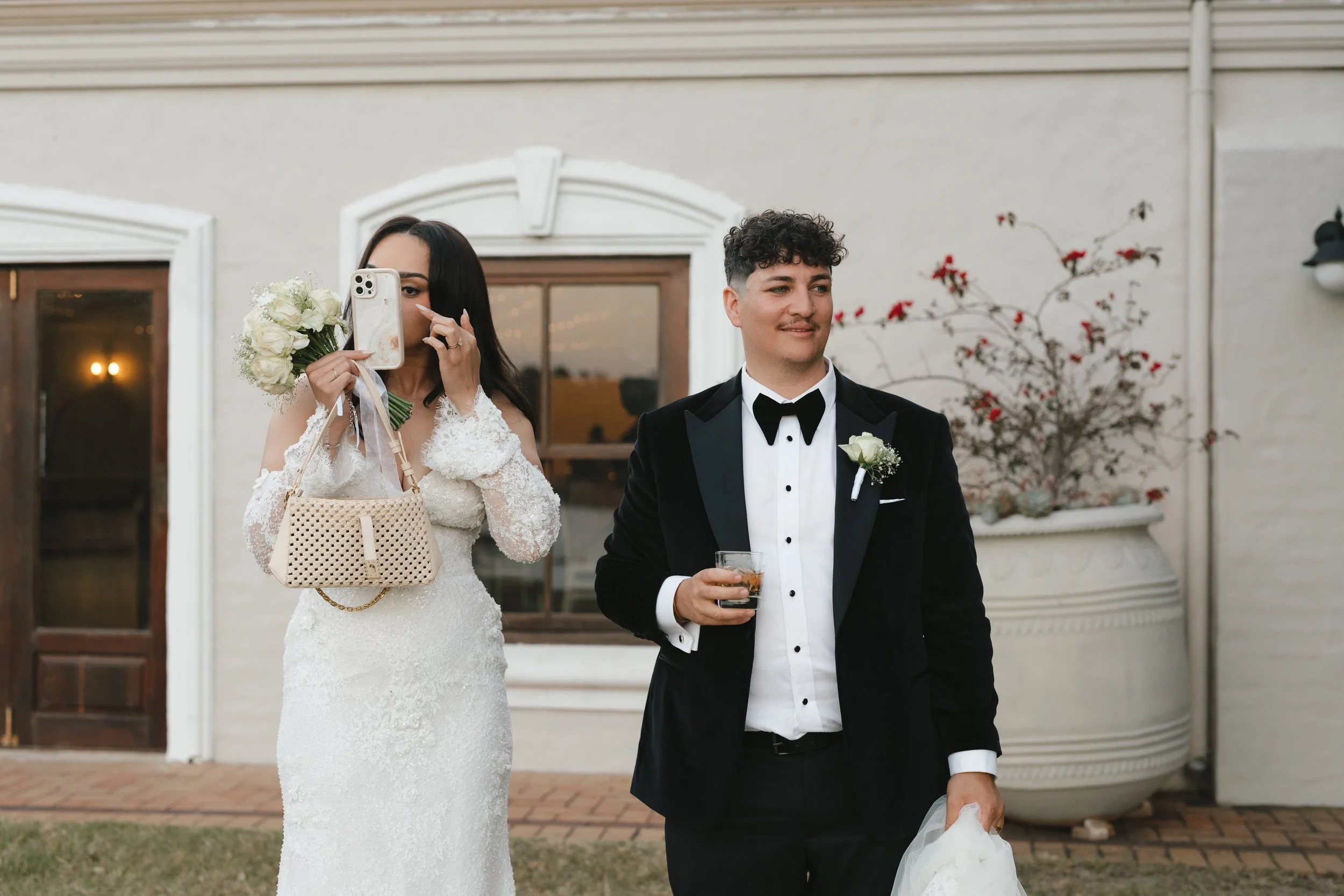 A bride and groom at their wedding, with the bride taking a photo and the groom holding a drink, standing outside a house. Gqeberha wedding photographer at Royalston Lakehouse, decor by The Hire Warehouse Flowers - Floral Creations