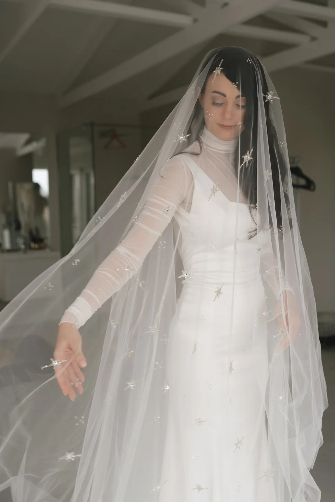 A woman in a white wedding dress with a sheer, long-sleeved top and a veil decorated with small embellishments, standing indoors with her eyes closed and a gentle smile. Gqeberha wedding photography at The Rose Barn, Jefferys Bay, dress by Werner Dey