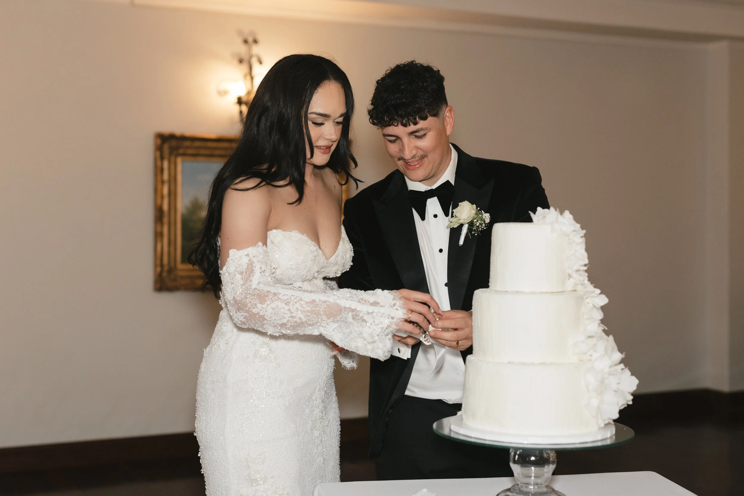 A bride and groom cutting their wedding cake together at a celebration. The bride is wearing a white lace wedding dress and the groom in a black tuxedo with a white boutonnière. Gqeberha wedding photographer Floral Creations