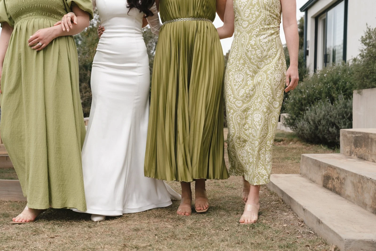 Four women standing outdoors, wearing dresses in shades of green and white, with gardening bushes and a house in the background. Gqeberha wedding photography at The Rose Barn, Jefferys Bay, dress by Werner Dey