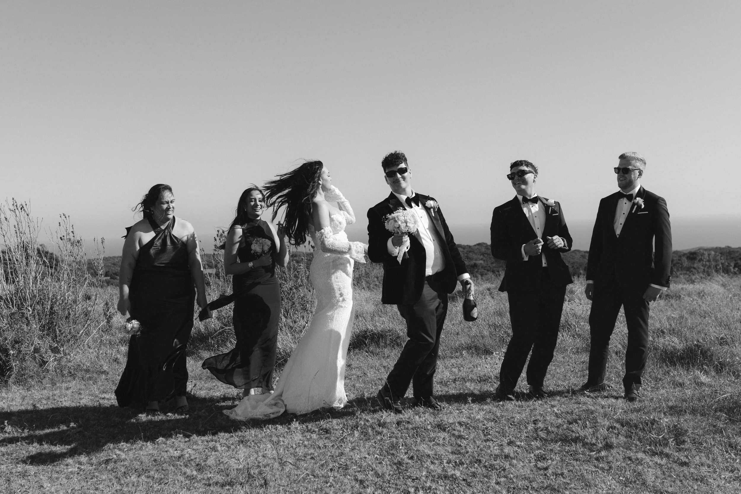 Black-and-white photo of a bride and five groomsmen walking outdoors on a sunny day, with clear sky and open landscape in the background. Gqeberha wedding photographer at Royalston Lakehouse.