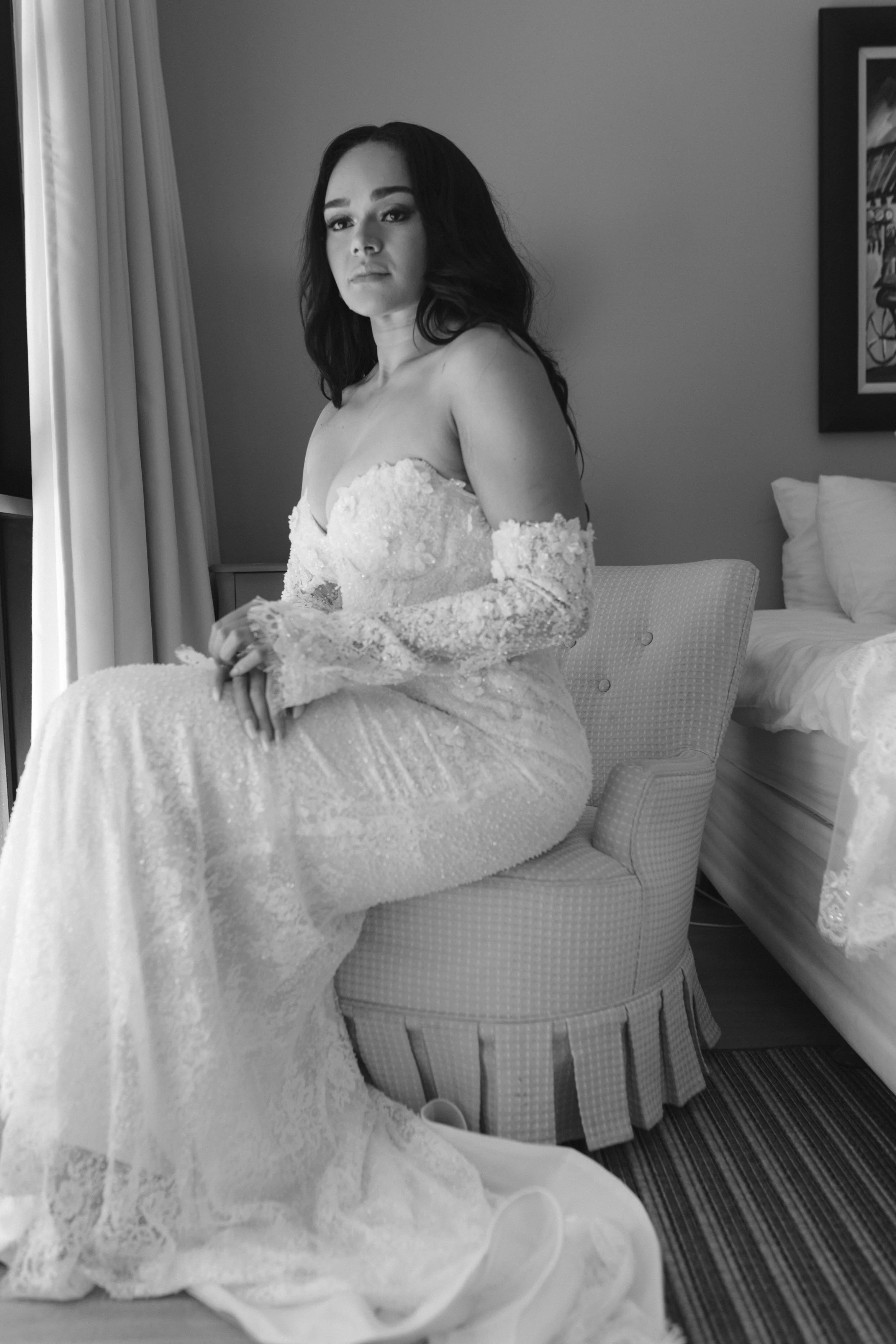 A woman in an off-shoulder lace dress sitting on a patterned chair in accommodation at royalston lakehouse, with a bed and artwork on the wall behind her. Gqeberha wedding photographer at Royalston Lakehouse.