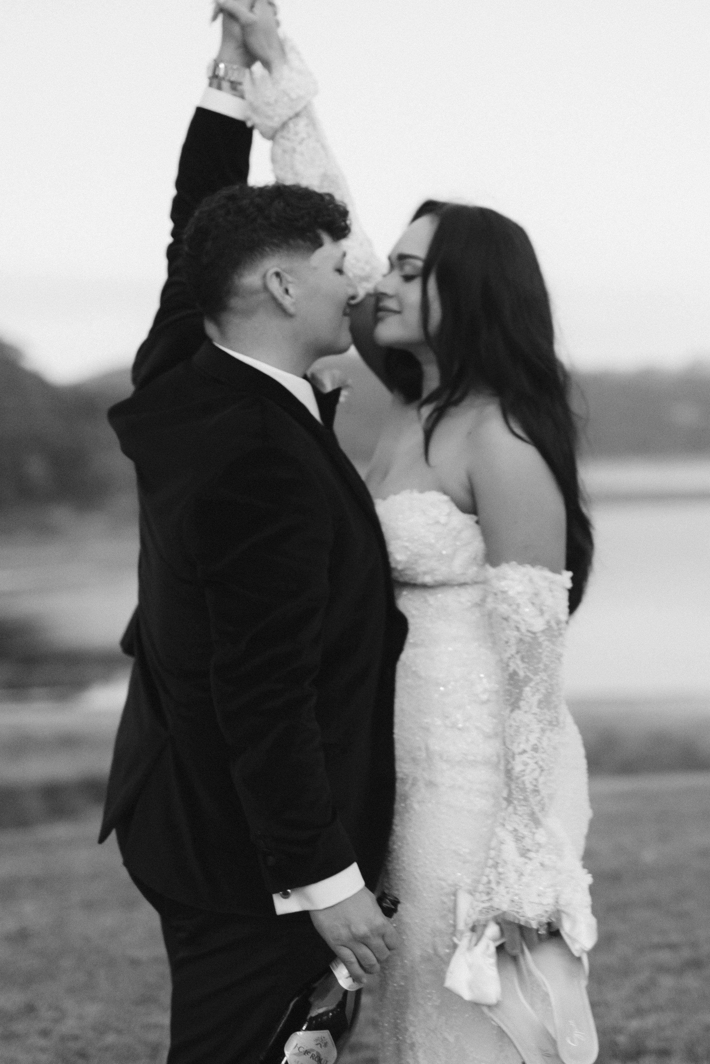 A black and white photo of a couple in wedding attire, standing close and touching foreheads, outdoors, holding hands, with their eyes closed and smiling softly. Gqeberha wedding photographer at Royalston Lakehouse, decor by The Hire Warehouse Flower