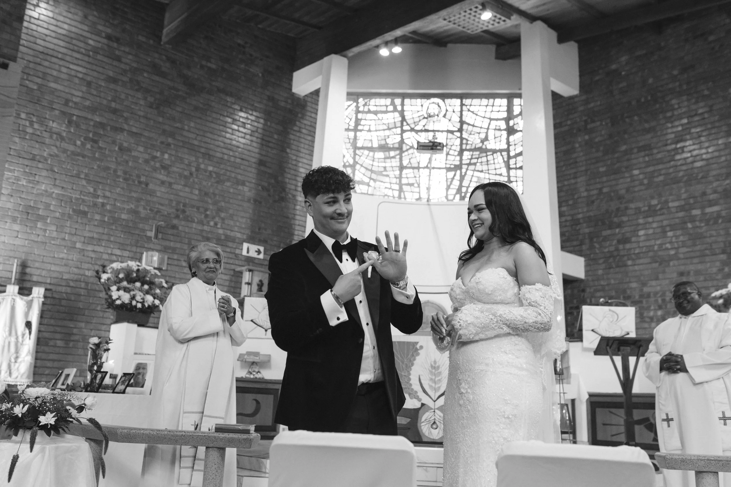 A wedding ceremony inside a church with a bride and groom exchanging rings, surrounded by a priest and guests, with stained glass windows and religious decor in the background. Gqeberha wedding photographer at Royalston Lakehouse.
