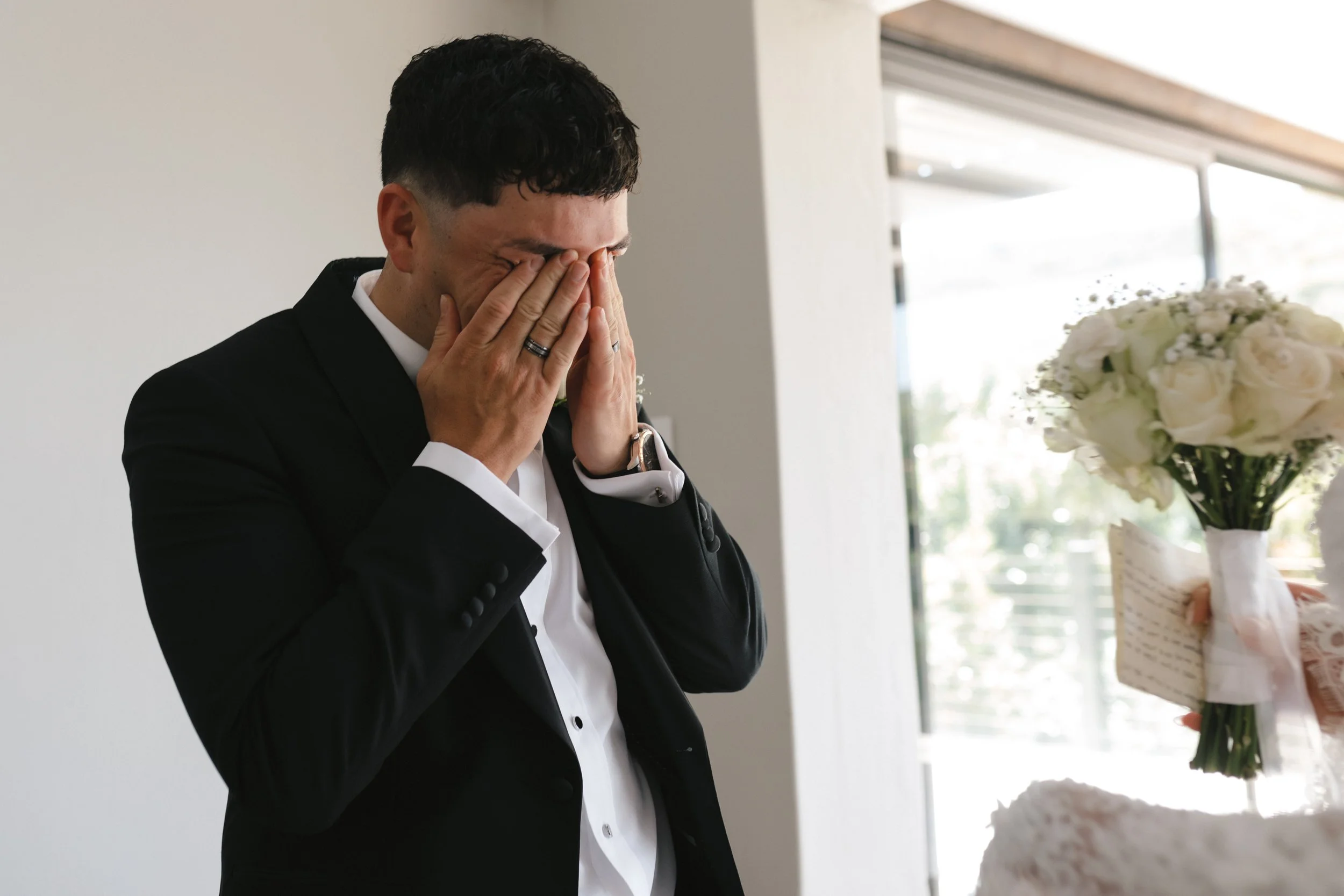 A man in a black suit with white shirt and rings cries with his hands while looking at the bride during daytime in a bright room. Gqeberha wedding photographer at Royalston Lakehouse.