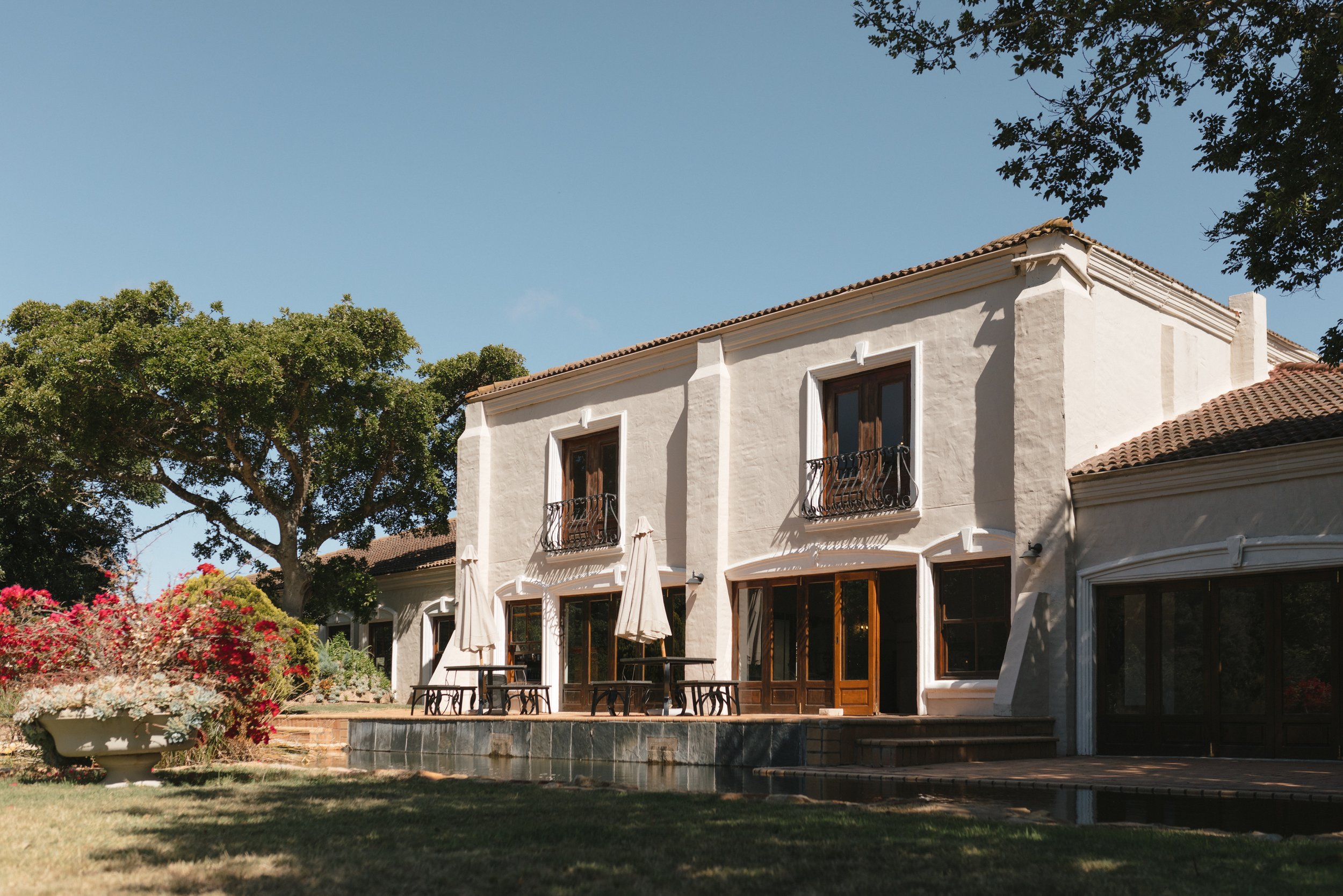 A white multi-story house with large wooden framed windows and French doors, surrounded by trees and colorful flowering bushes under a clear blue sky. Gqeberha wedding photographer at The Plantation Wedding Venue.