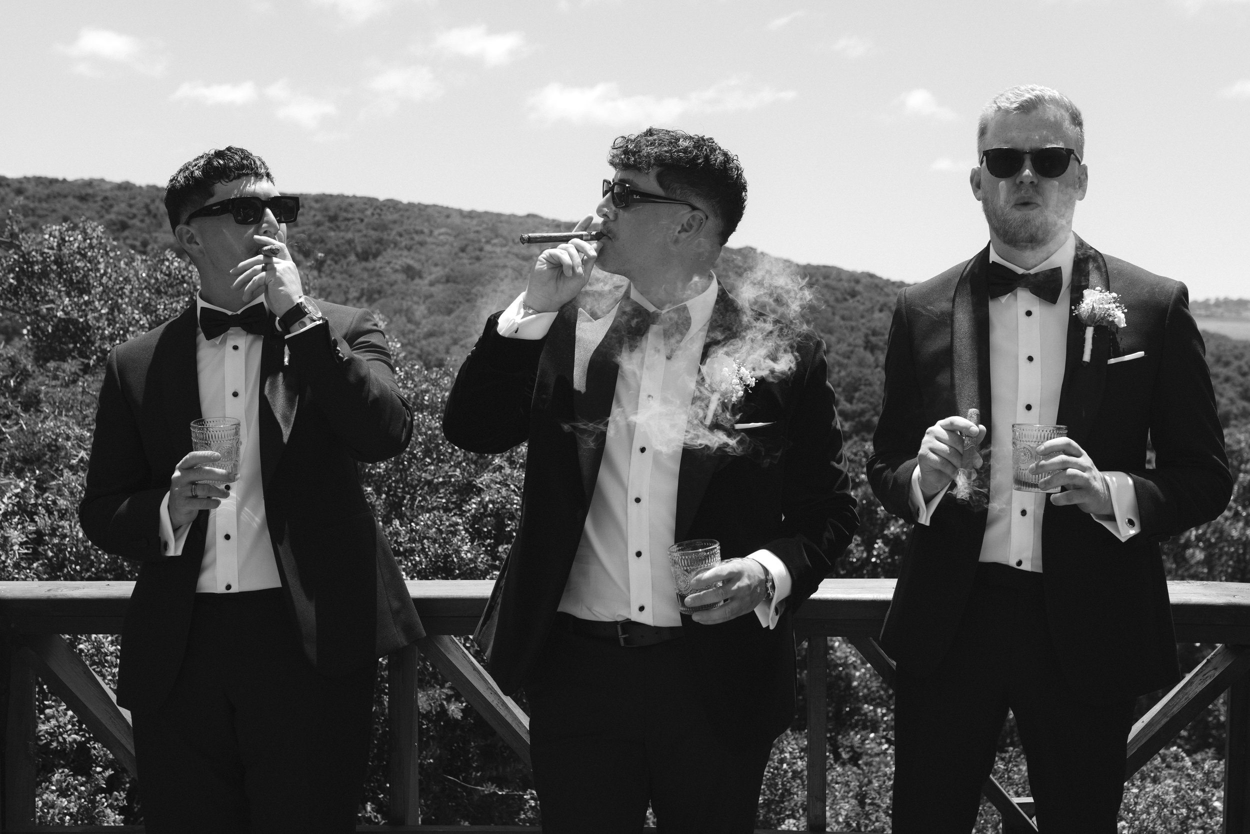Three men in tuxedos and sunglasses standing outdoors, smoking cigars, holding glasses, with a natural landscape and blue sky in the background. Gqeberha wedding photographer at Royalston Lakehouse.